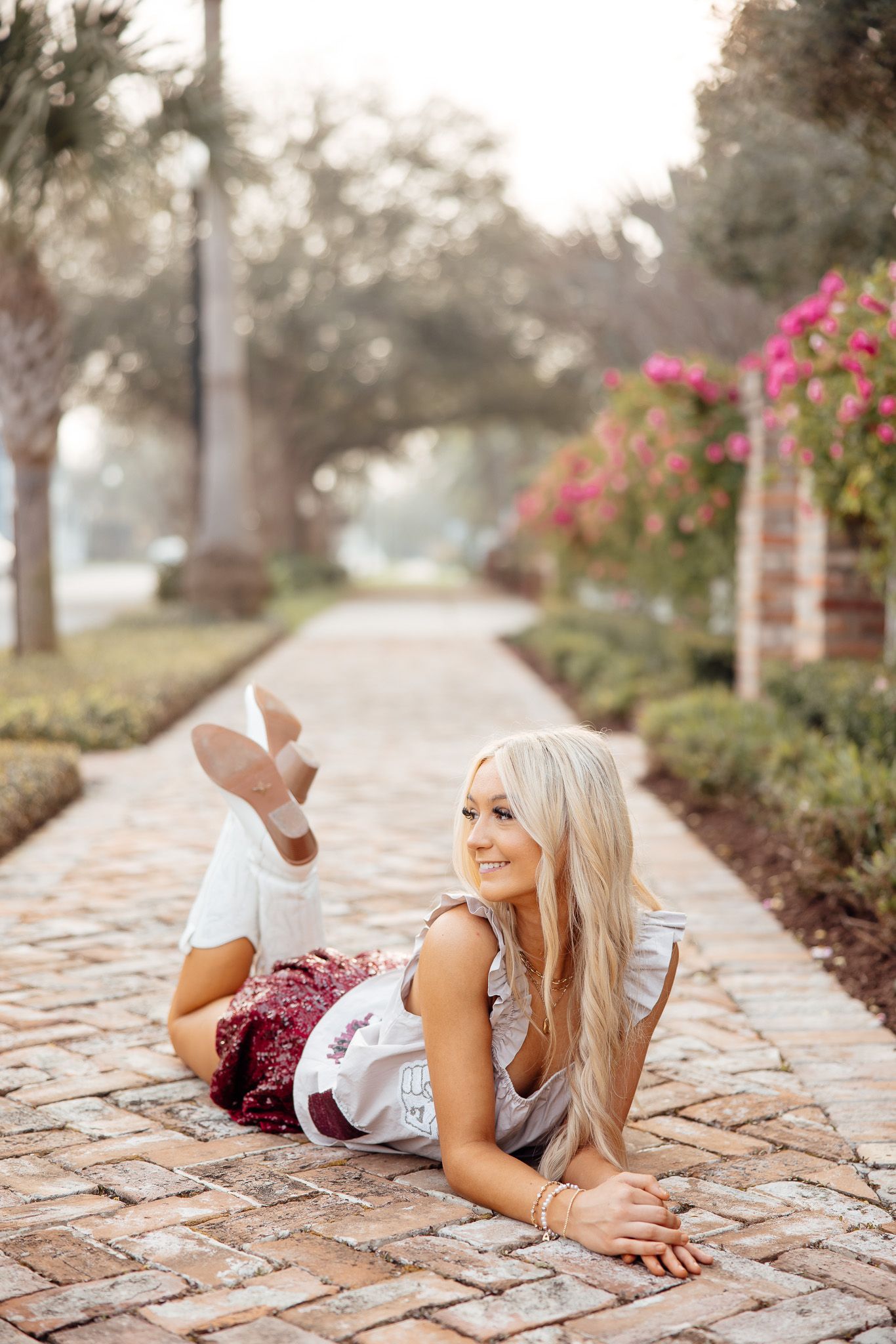 A person in a maroon cheerleading uniform poses on a brick path surrounded by pink flowers and greenery.