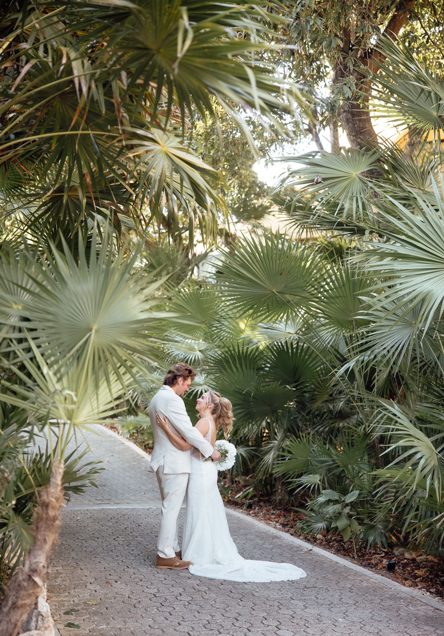 Romantic wedding couple embraces on a path surrounded by tropical palm fronds in a garden setting.