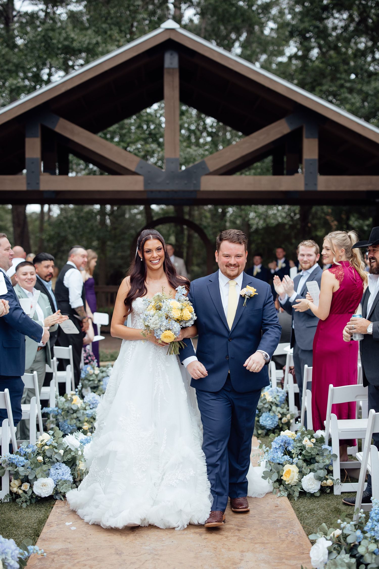 Bride and groom walk down the aisle after outdoor wedding ceremony under wooden pavilion surrounded by blue and white floral arrangements.