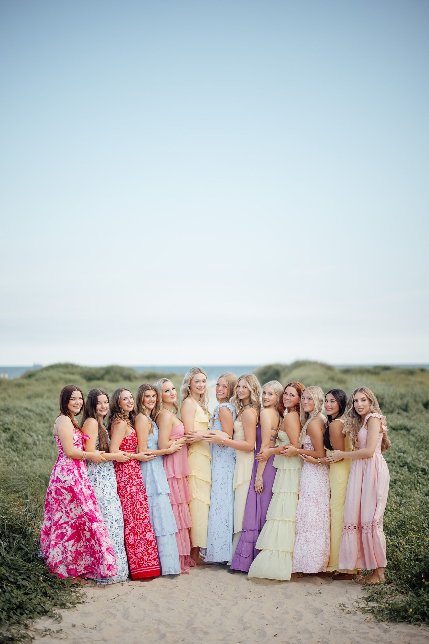 A group in colorful pastel tiered maxi dresses pose together on a sandy path with grassy dunes in the background.
