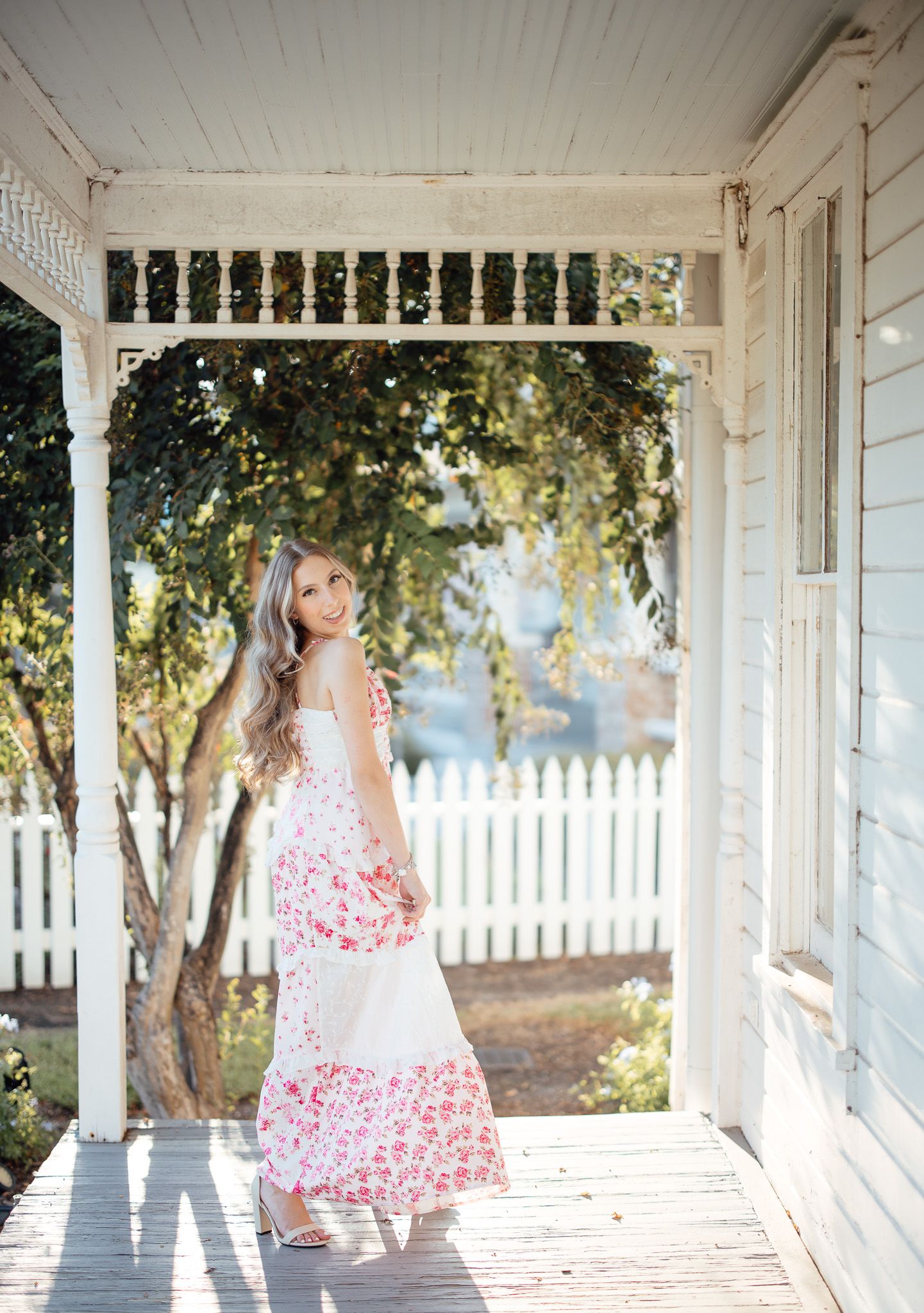 A woman in a pink floral dress poses on a white wraparound porch with sunlit trees in the background.