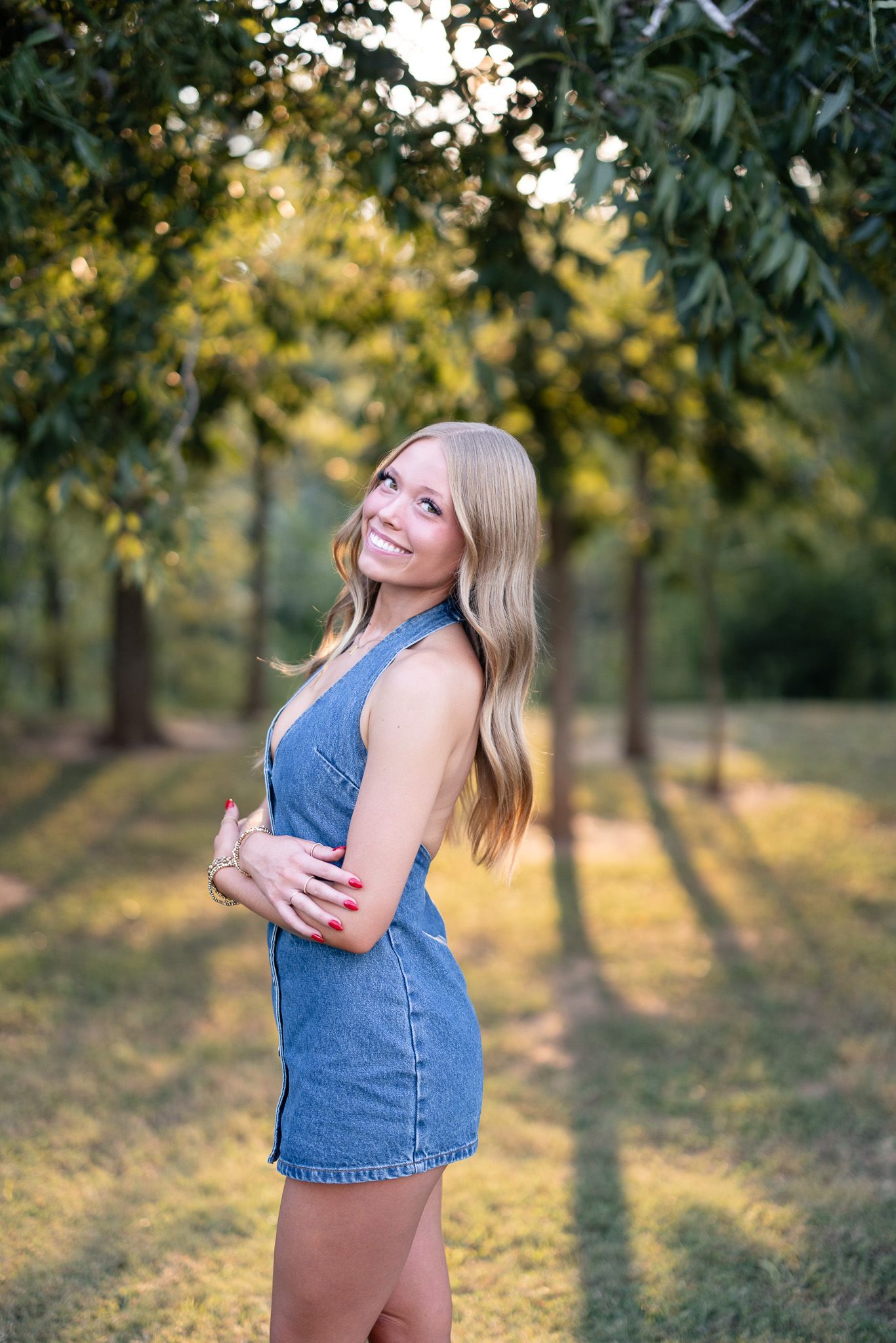 A person in a denim top poses playfully in a sun-dappled orchard during golden hour.