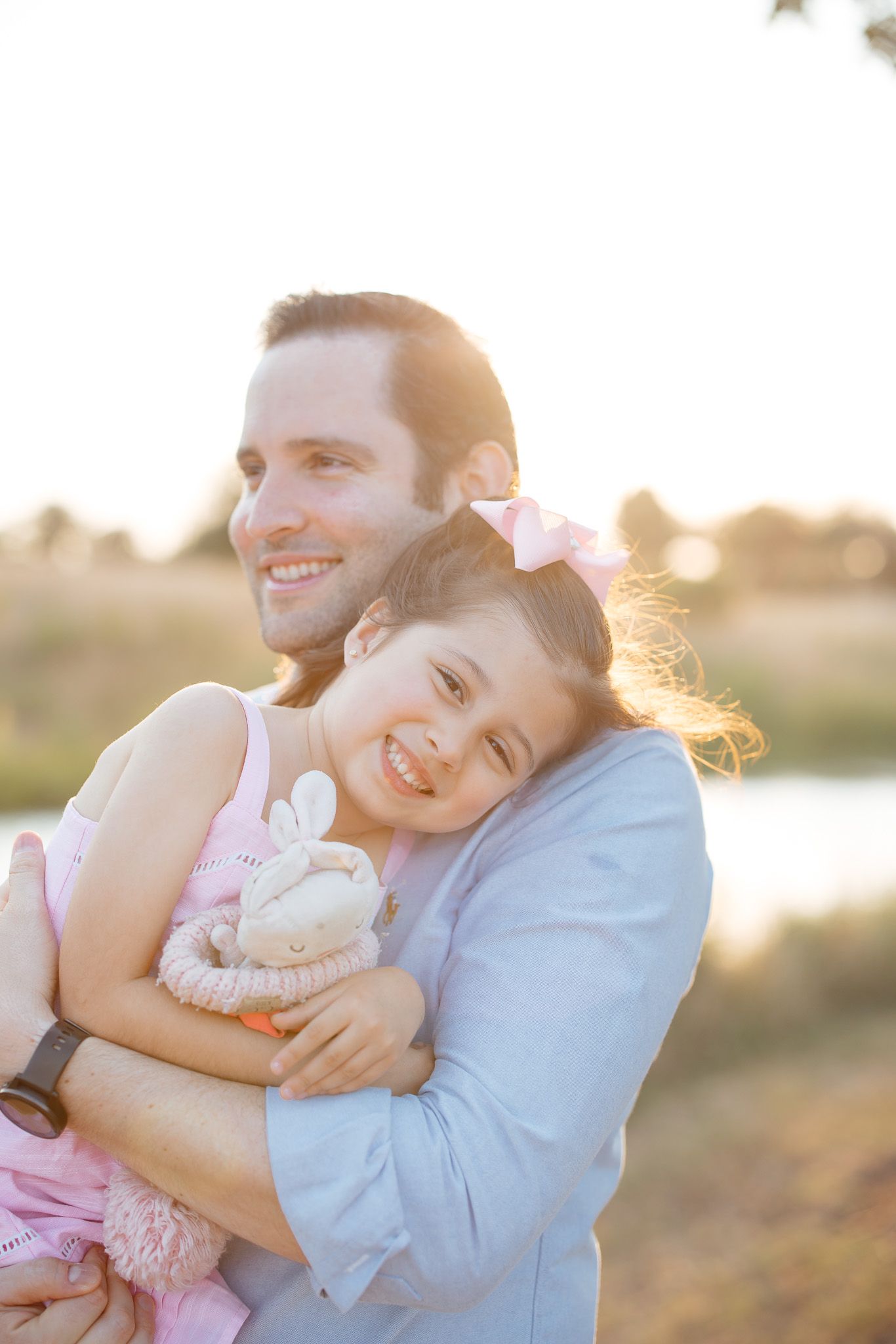 A parent and child share a joyful embrace while holding a stuffed toy during golden hour outdoors.