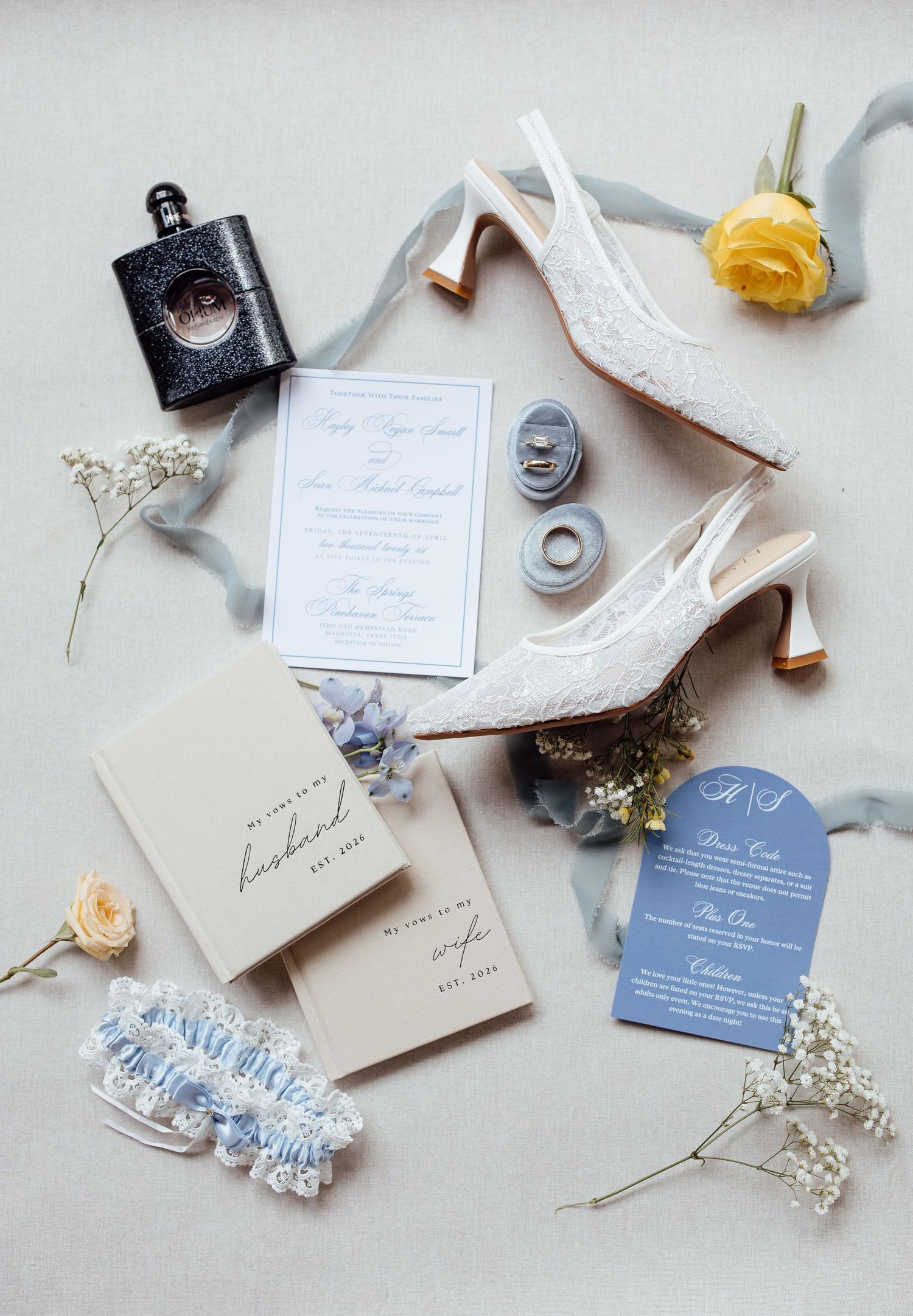 Elegant wedding flat lay with lace heels, floral invitation, blue garter, rings, perfume, and soft florals on white background.