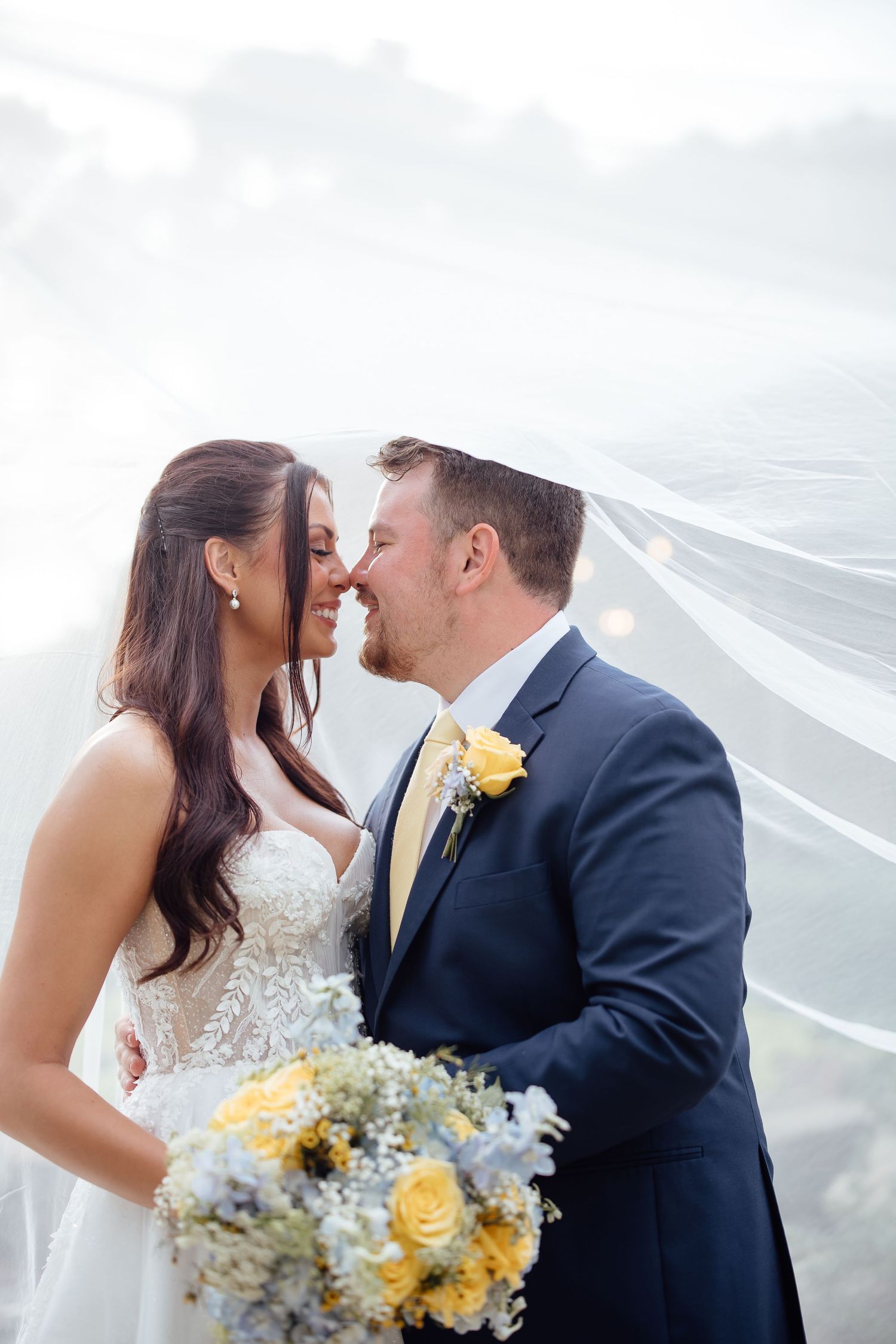 Bride and groom sharing a romantic moment under a flowing veil, holding a yellow and blue bouquet.
