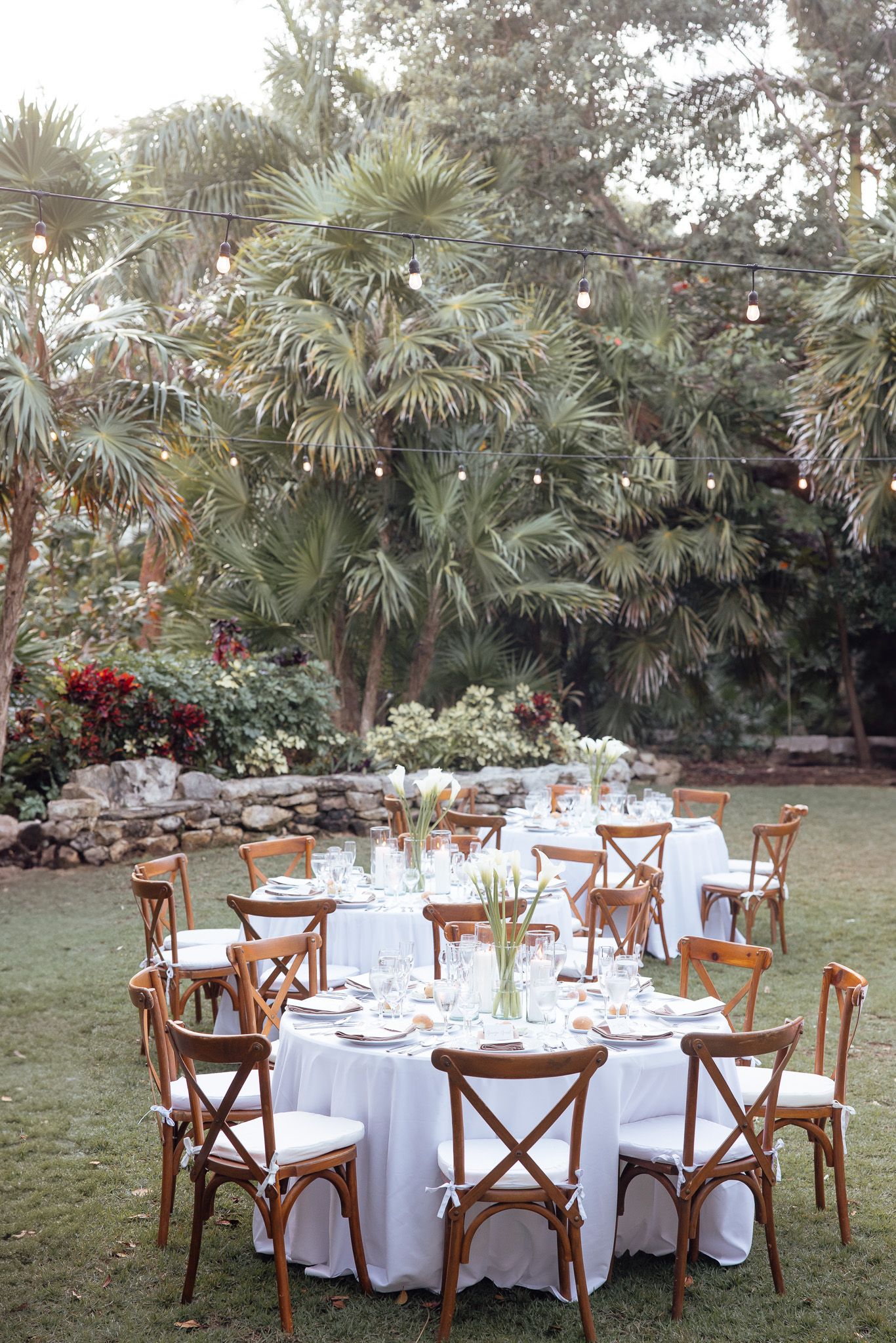 Elegant outdoor wedding reception setup with round tables, white linens and wooden cross-back chairs under tropical palm trees.