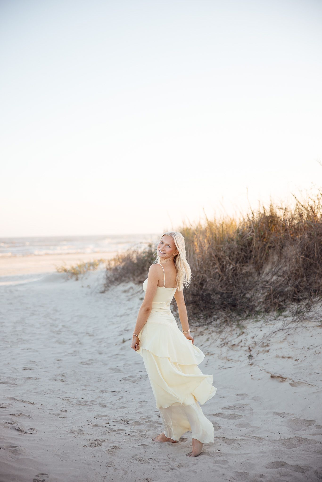 A person in a flowing yellow dress walks barefoot along a misty beach at sunset.