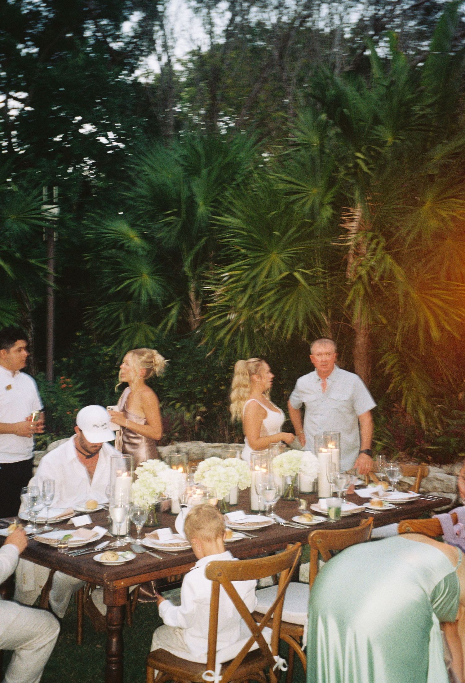Elegant outdoor dinner party setup with long tables and white linens under palm trees.