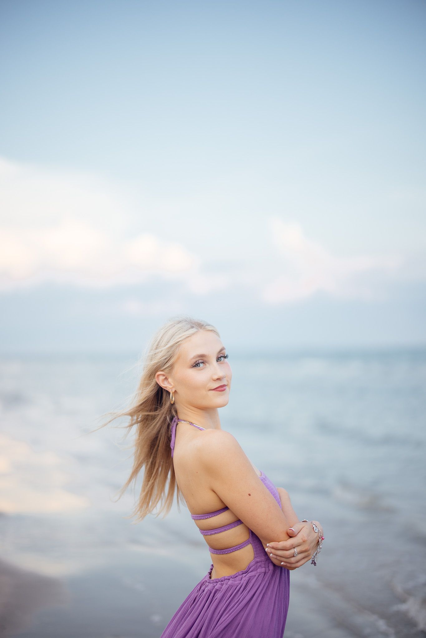 A person in a purple dress poses dreamily by the ocean at dusk with soft lighting and blurred background.
