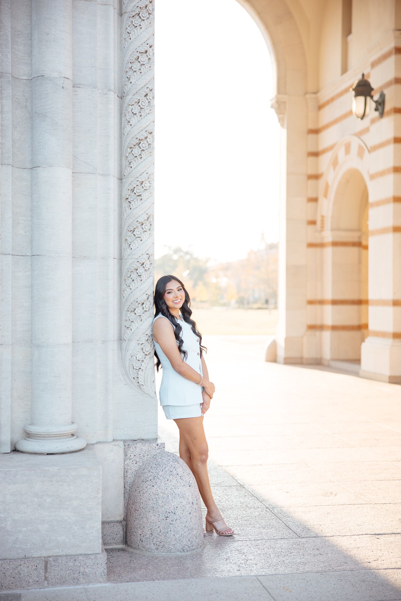 A person in a white dress poses gracefully against white stone columns in warm sunlight.