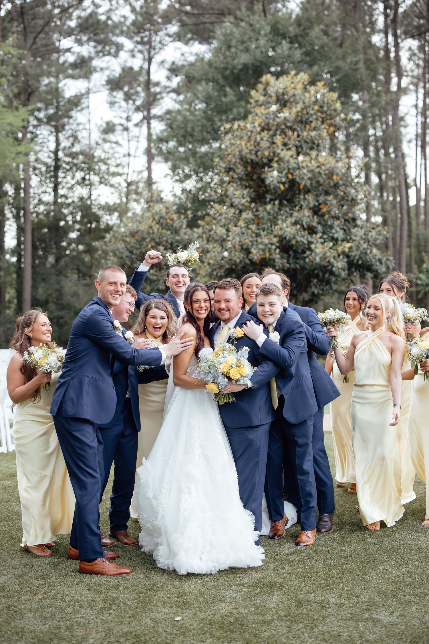 Wedding party in navy suits and yellow gowns celebrating with bride and groom outdoors on green lawn with trees behind.