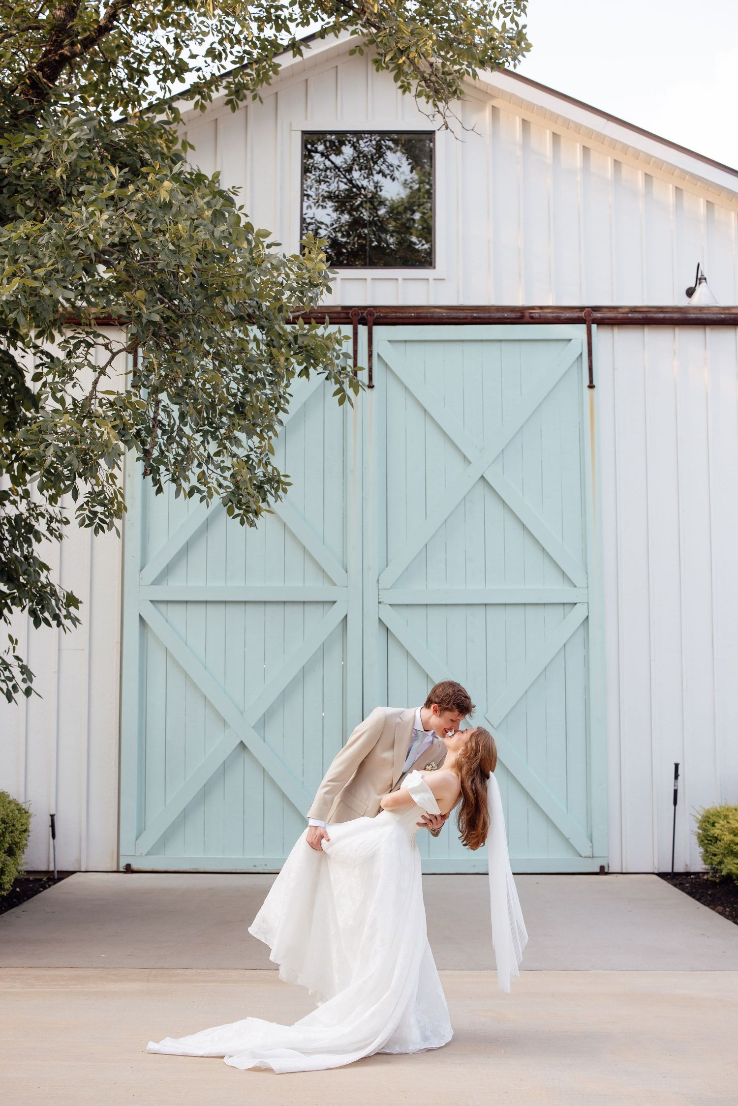 Bride and groom share a romantic dip kiss in front of white barn with mint green doors at outdoor wedding venue.