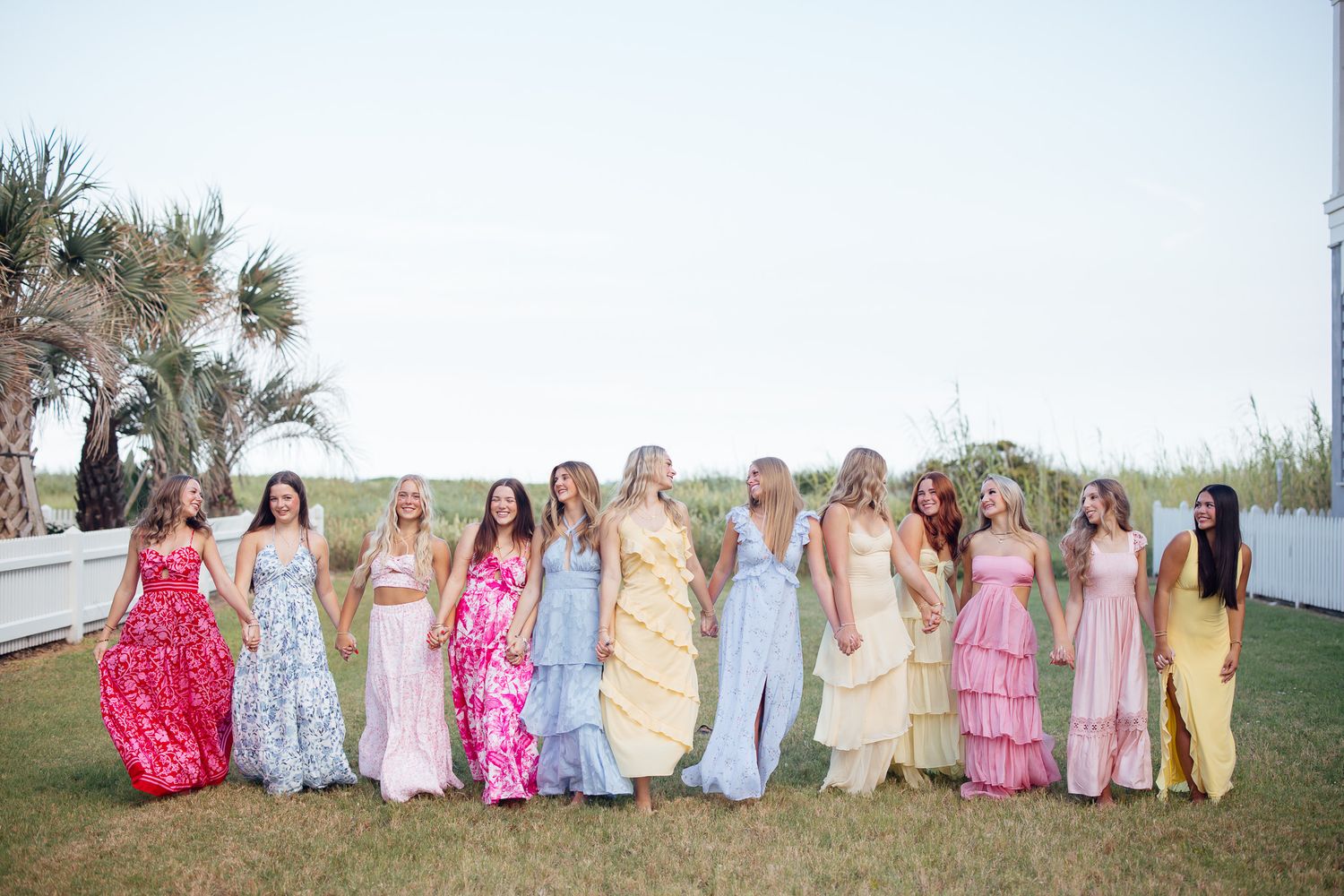 A group in colorful formal dresses and gowns stands together in a line against a beachside backdrop.