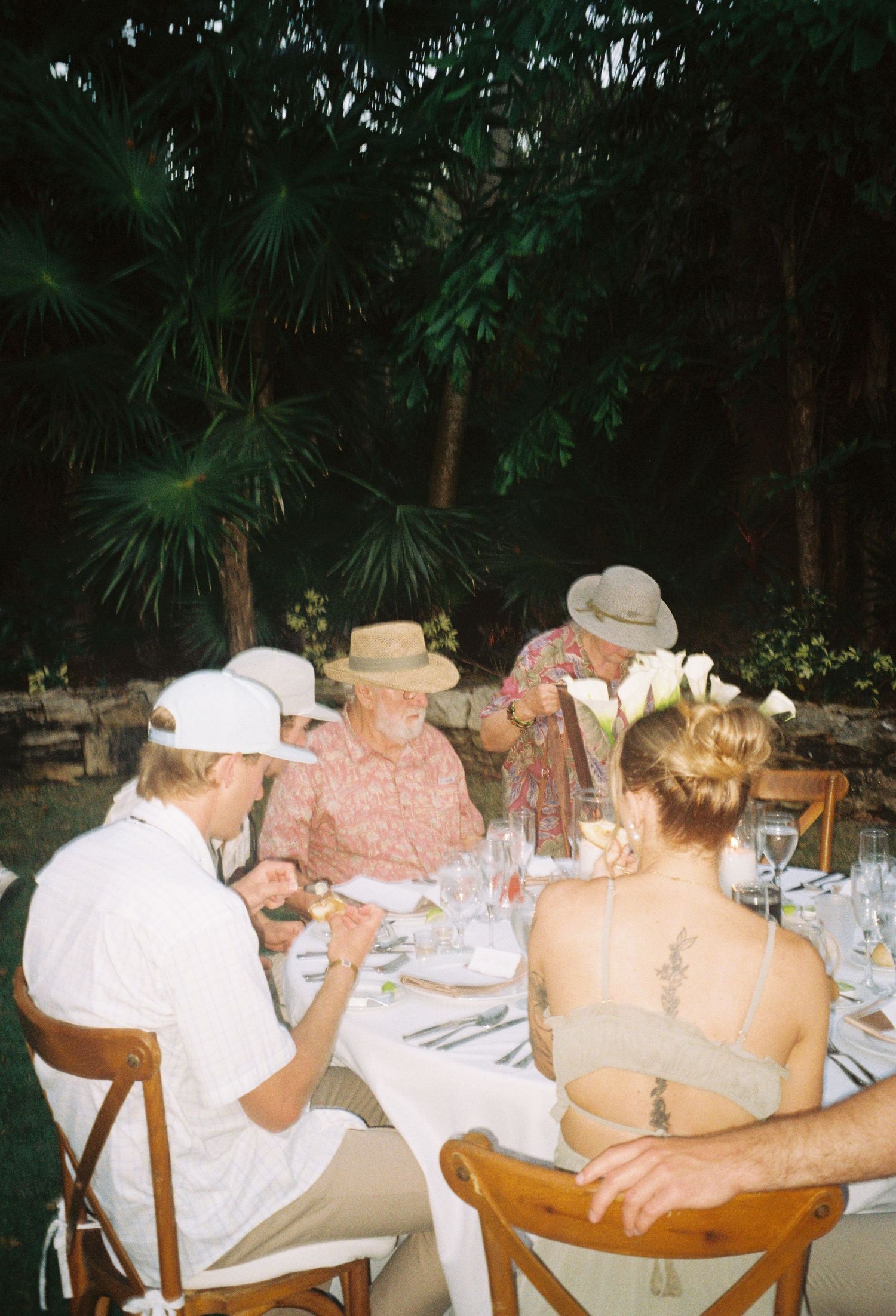 Guests seated at an outdoor dinner table surrounded by lush tropical foliage during evening hours.
