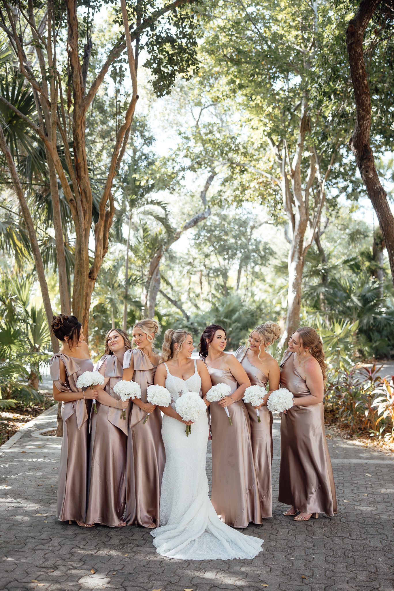 Wedding party in champagne-colored gowns poses together on a garden path surrounded by lush tropical trees.