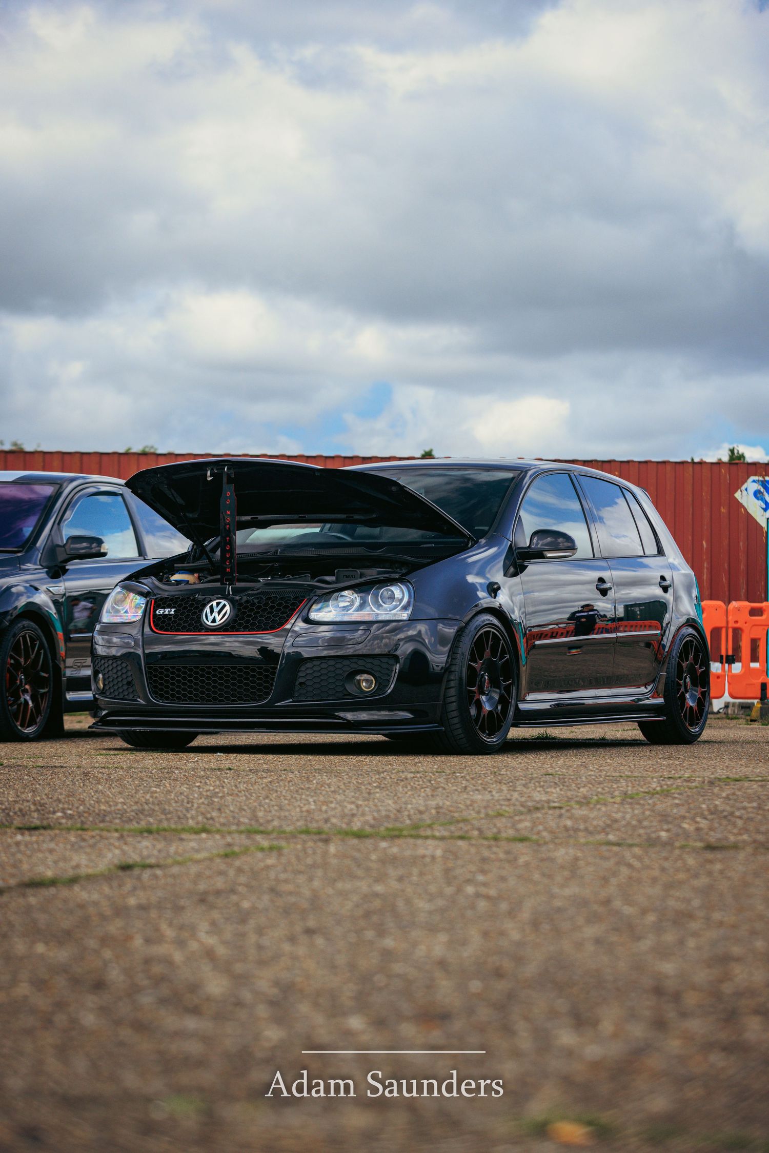 A lowered black Volkswagen GTI with black wheels parked on dirt ground under cloudy skies.