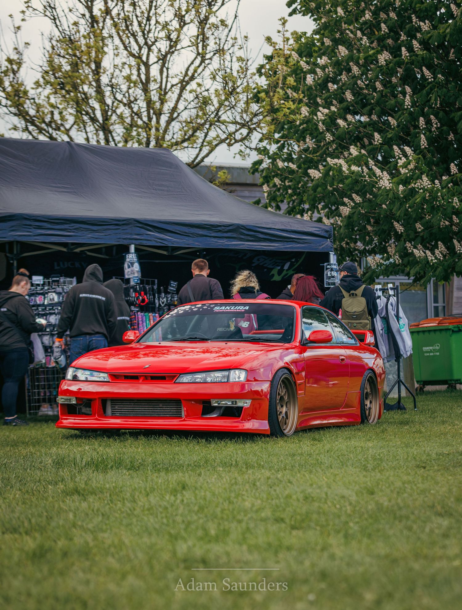 A lowered red Nissan Silvia S14 sports car displayed at an outdoor car show with vendor tents in the background.