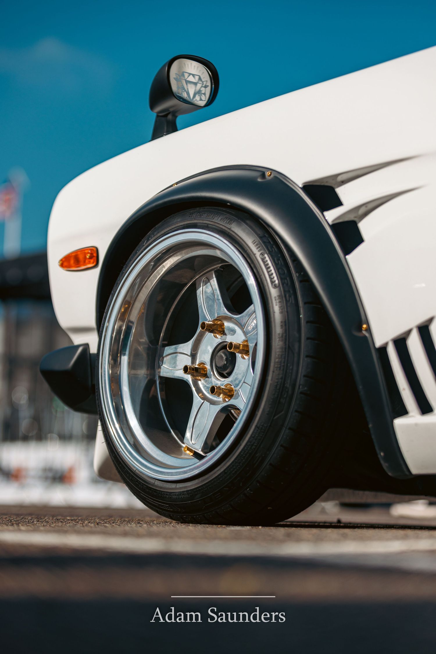 Low angle automotive photo showing white fender and polished silver sports wheel on a classic car against blue sky.