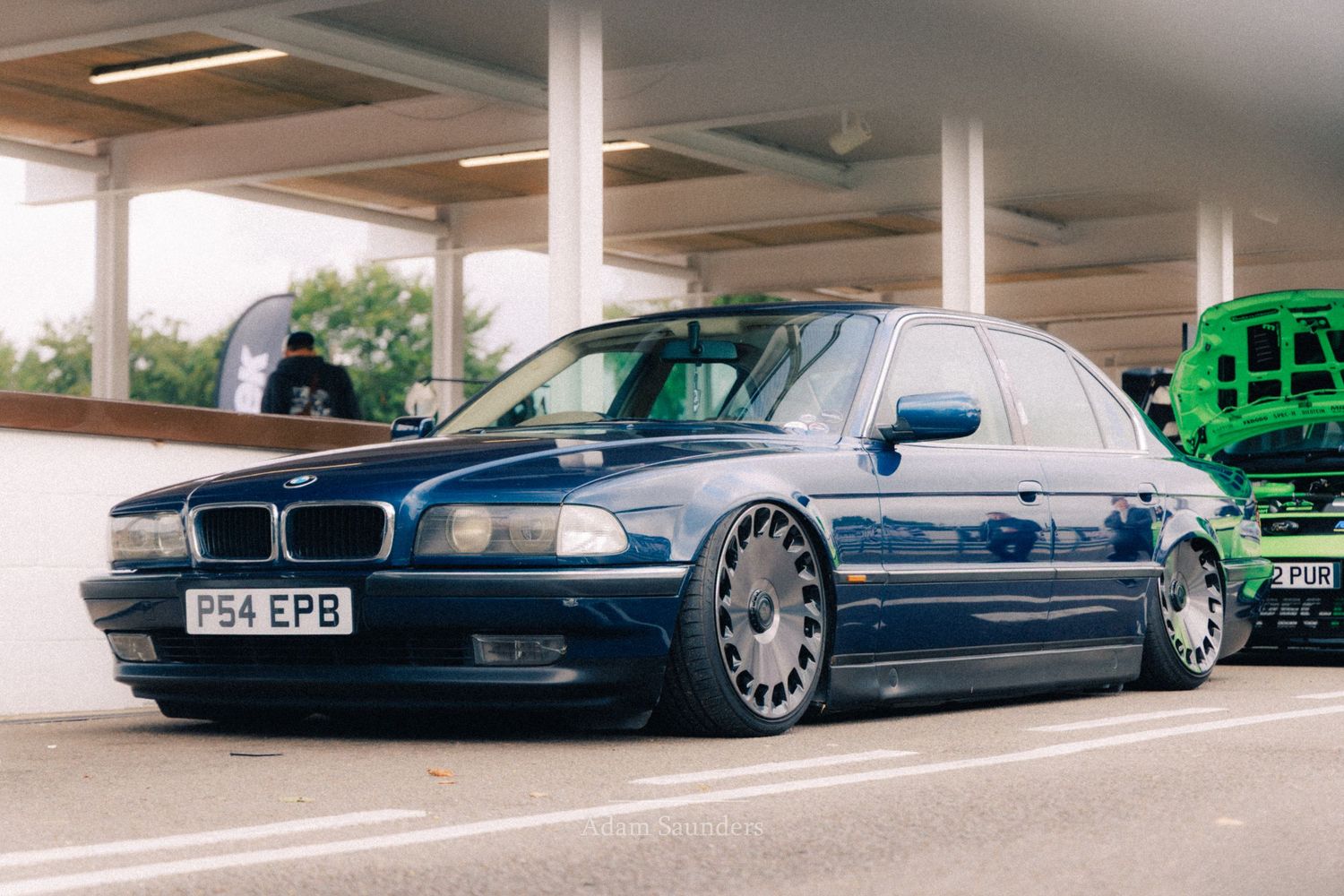 A lowered navy blue BMW E38 7 Series sedan on custom wheels, parked in a car park at a meet.