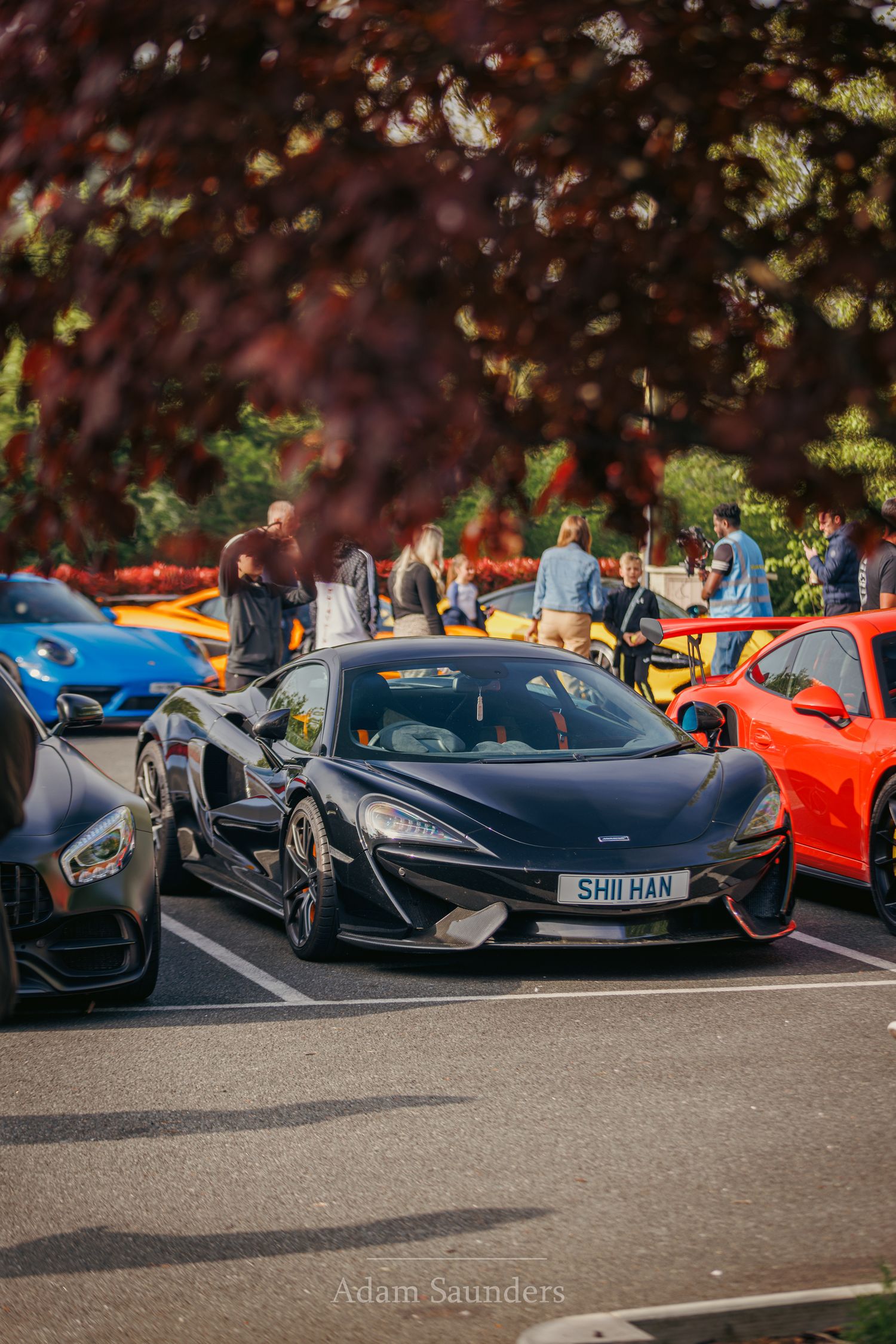 A sleek McLaren sports car in metallic black sits among other luxury vehicles at a car show with autumn foliage in the background.