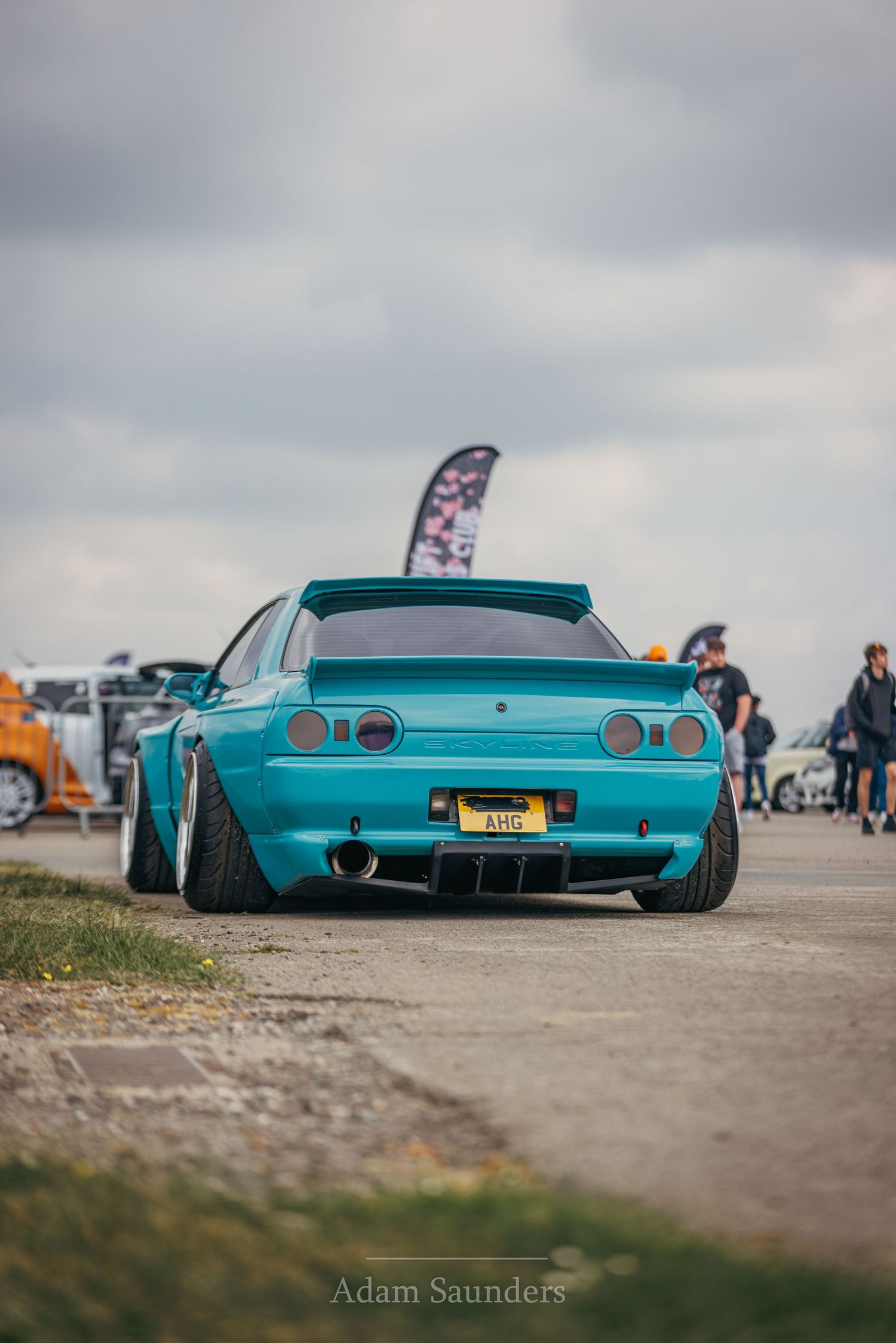 Low-stance turquoise Nissan Skyline with custom bodywork and wheels parked at a car show under cloudy skies.