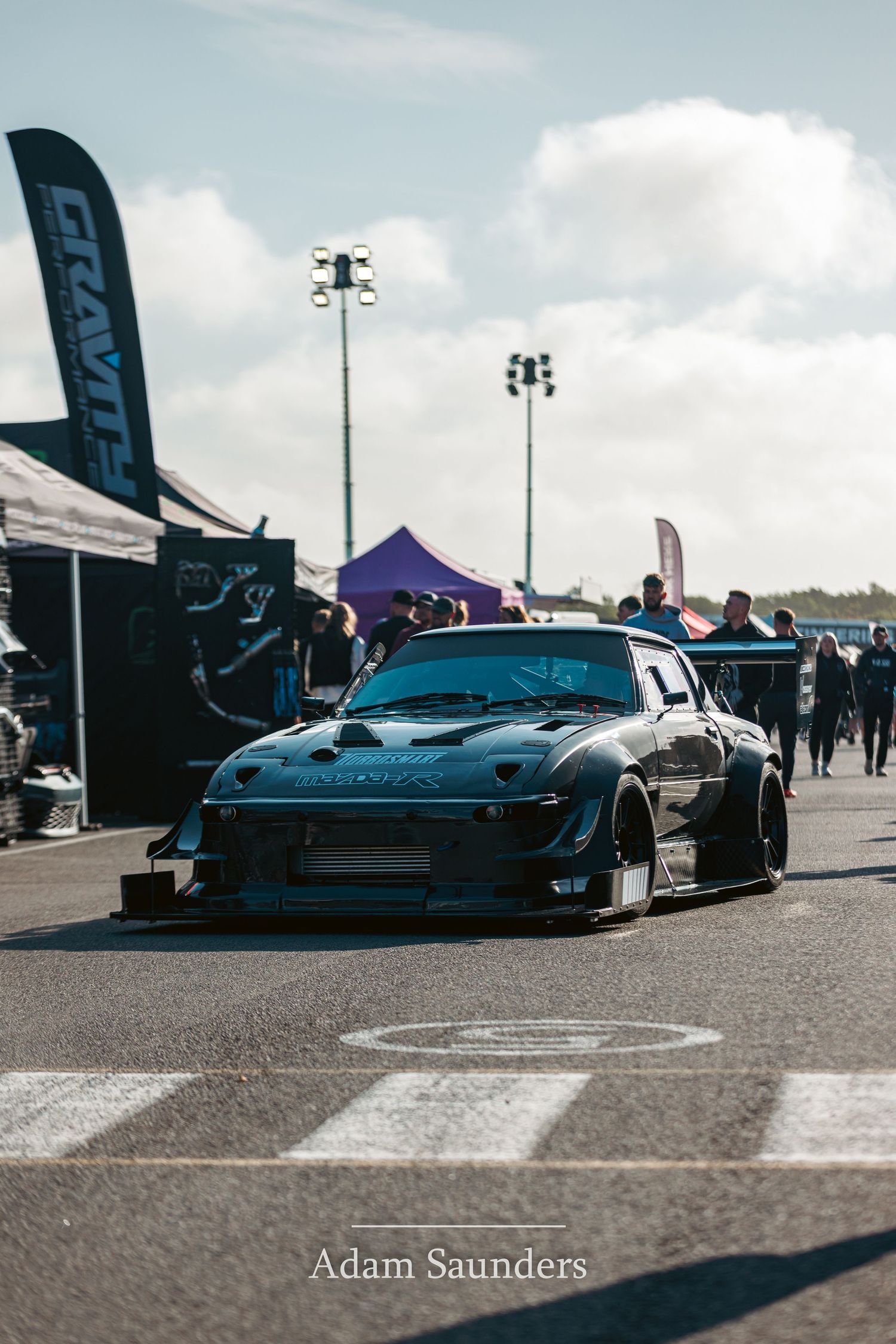 Modified sports car with wide body kit and blue accents parked at outdoor motorsports event with tents and light poles.