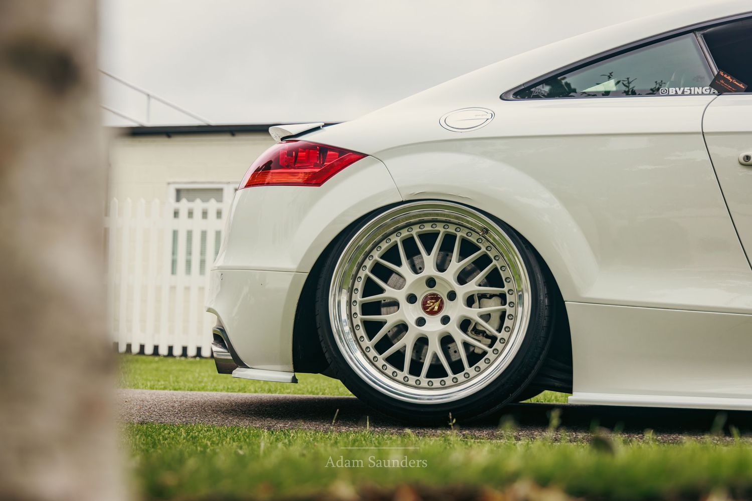 White Audi TT coupe with polished BBS-style alloy wheels, slammed stance, parked on driveway near white fence.