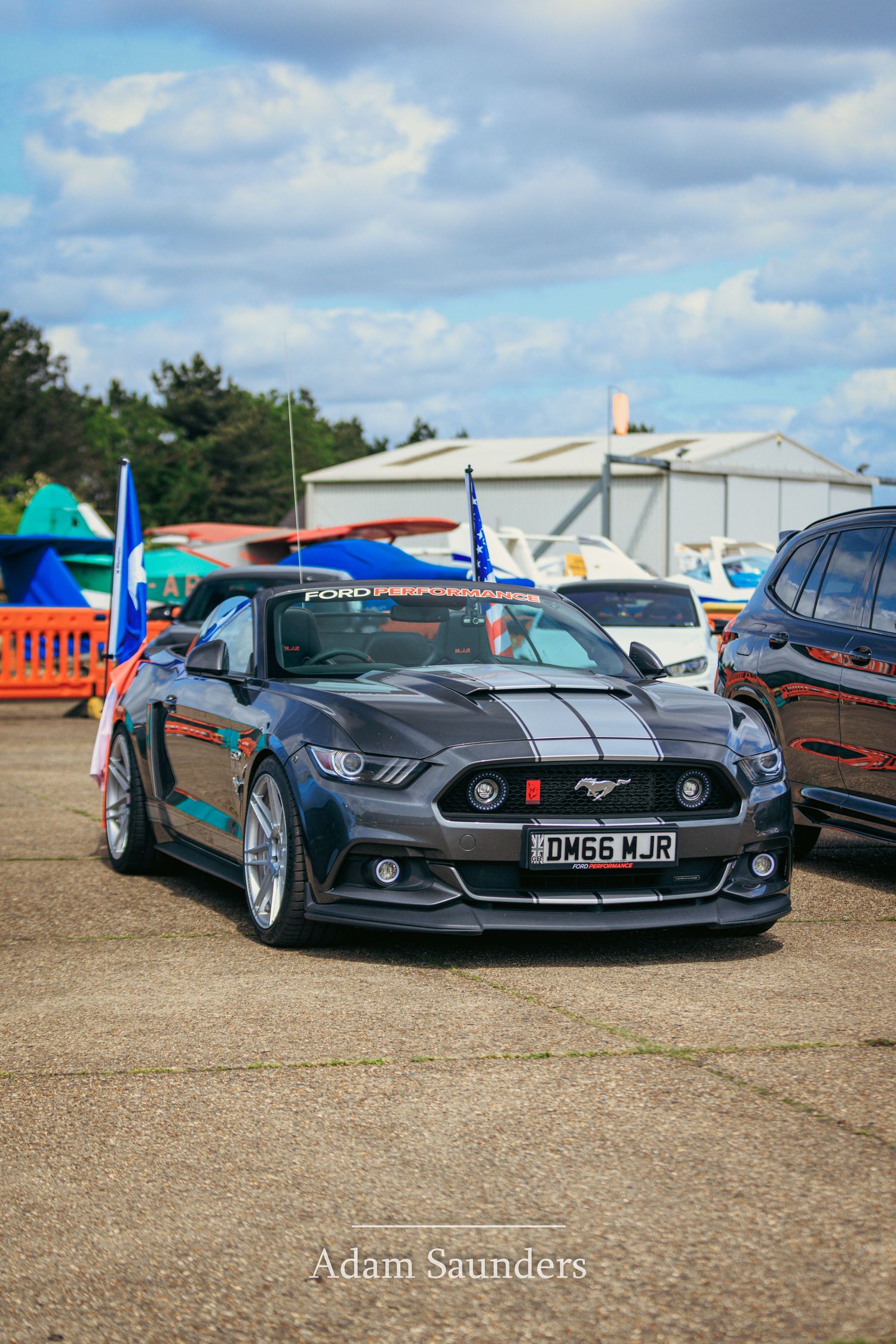 A modified gray Ford Mustang with black racing stripes and custom wheels parked at an outdoor car show.