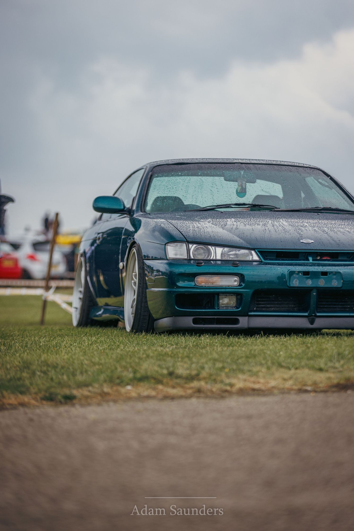 Modified teal Nissan Silvia with lowered suspension and aftermarket wheels on grass.