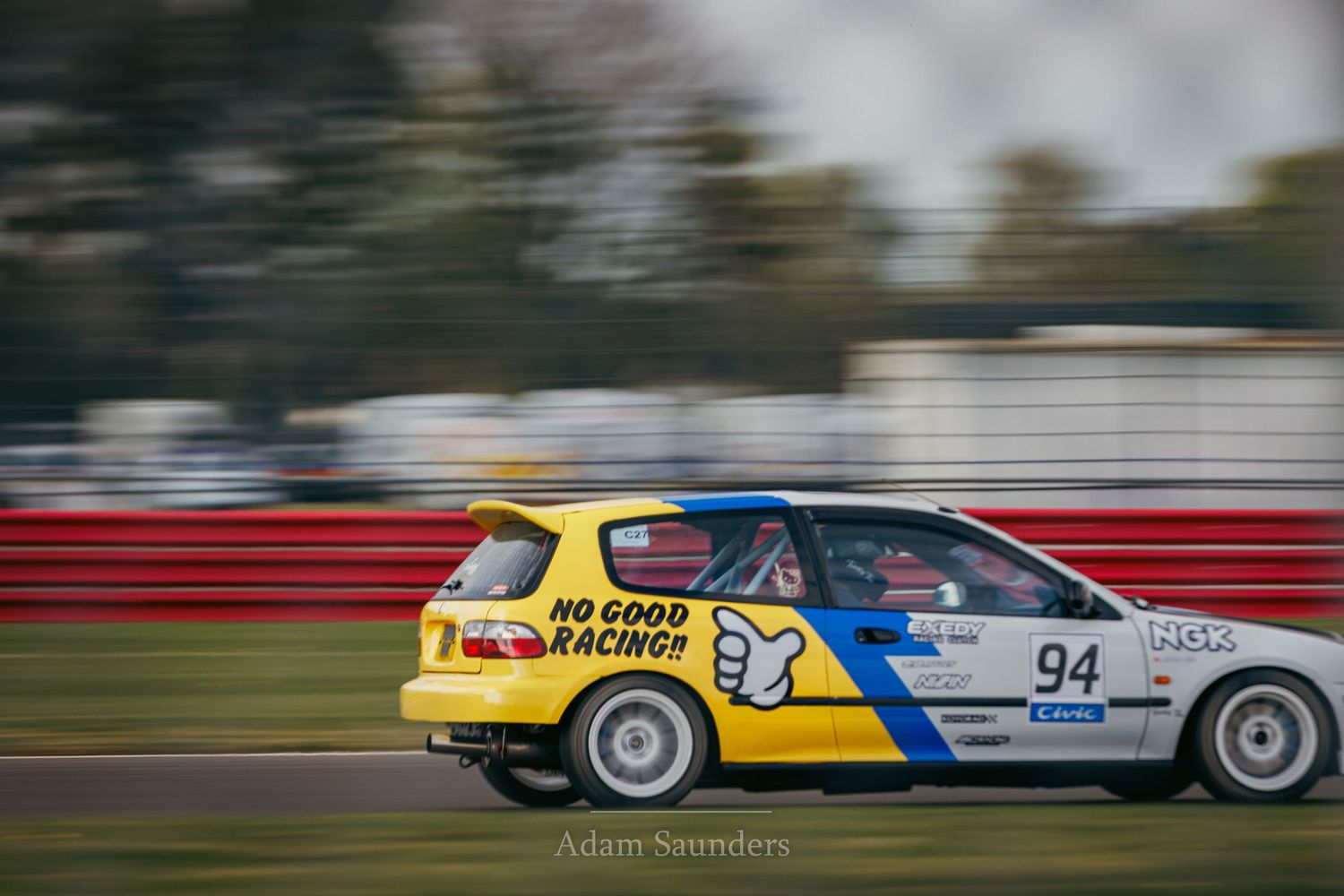 A race-spec Honda Civic hatchback with racing livery speeds past on a racetrack with motion blur effect.
