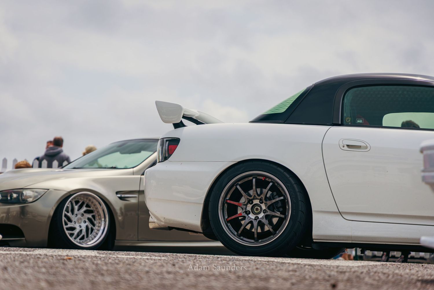 Two lowered sports cars, a white Honda S2000 and gold BMW, parked on a track with stylish aftermarket wheels.