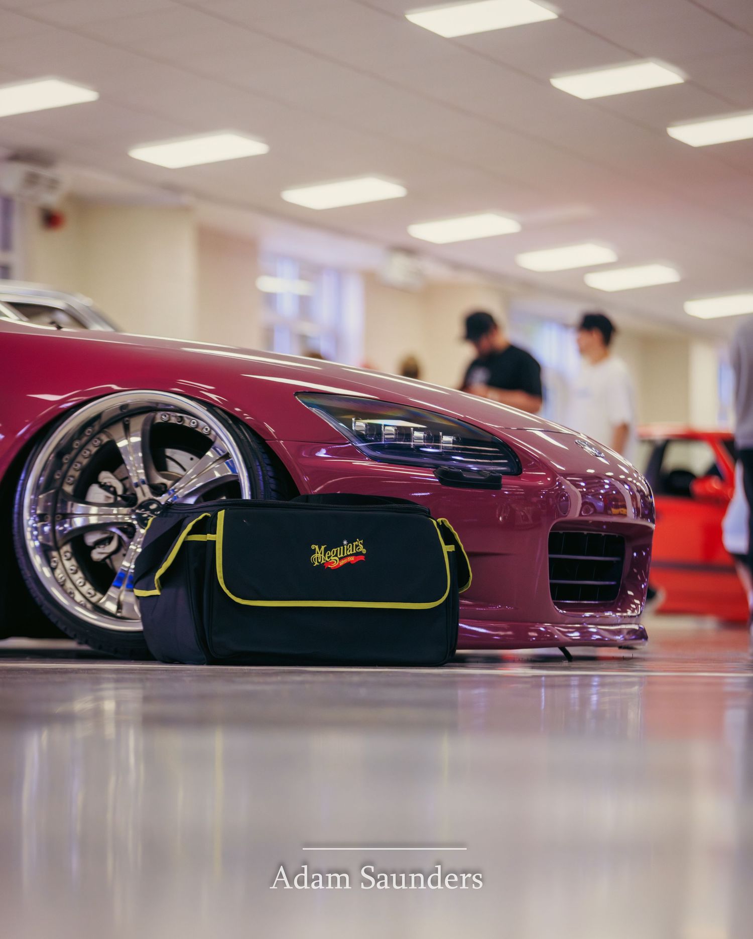 A burgundy sports car with chrome wheels sits next to a black Meguiar's detailing bag in a well-lit showroom.