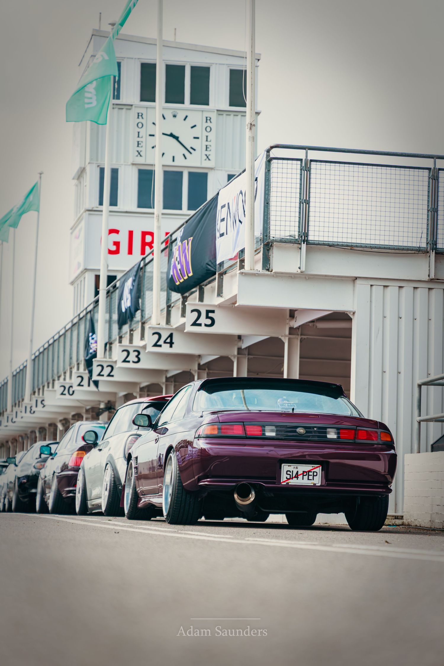 Purple sports car parked in pit lane at Goodwood Circuit, with Rolex timing tower and race flags visible in background.