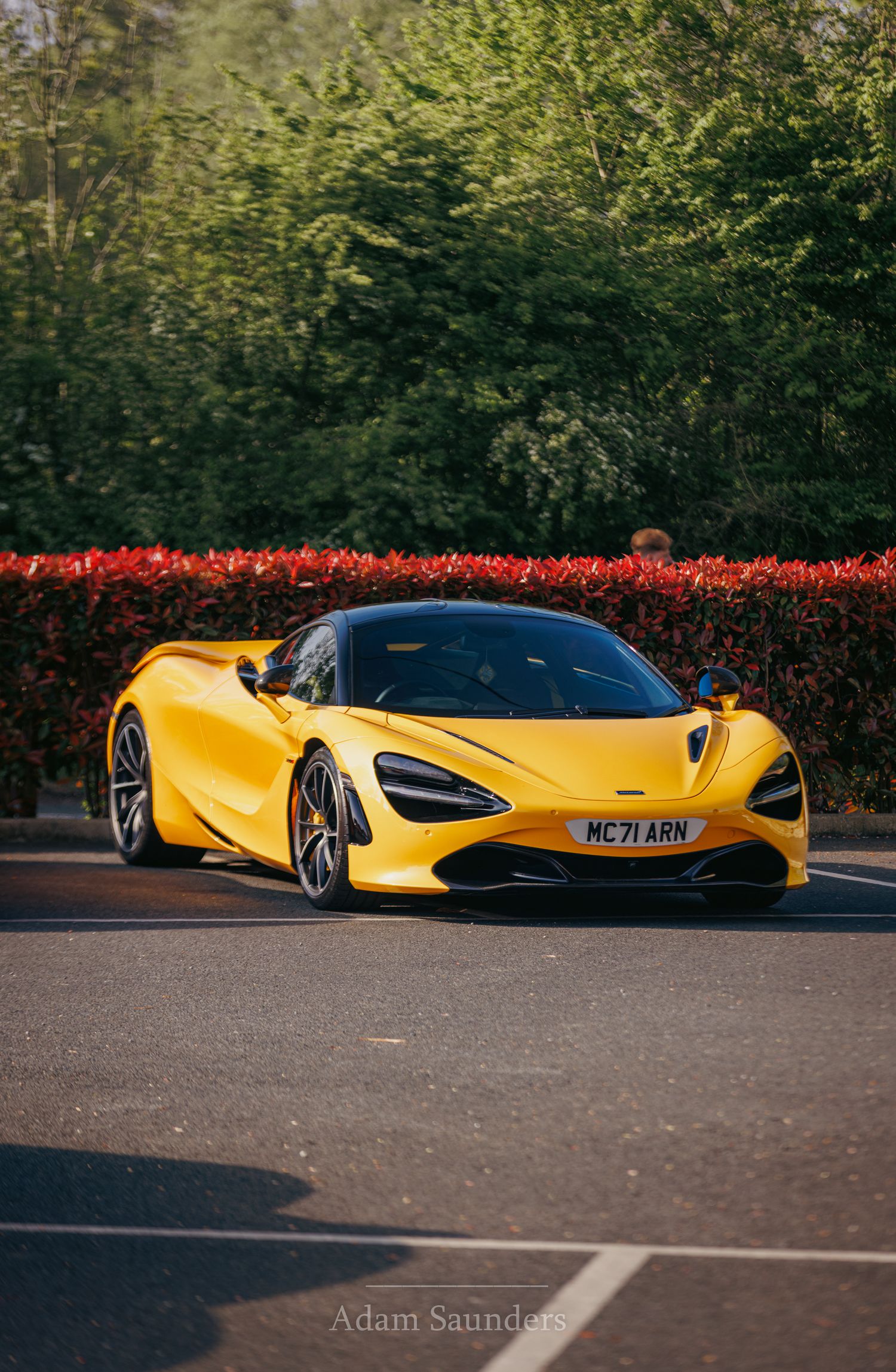 A bright yellow McLaren 720S supercar parked in front of red hedges and green trees on a sunny day.