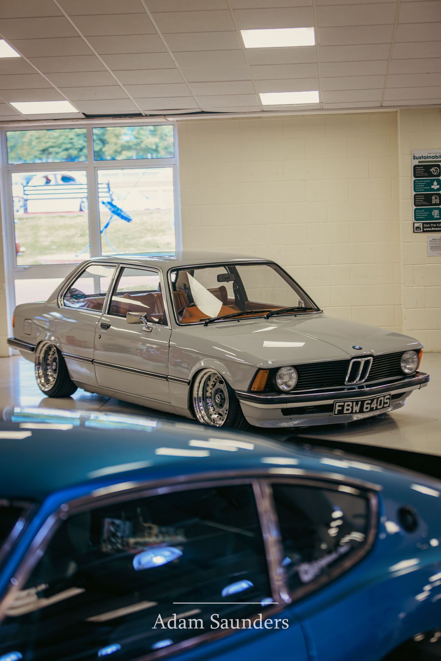 A lowered silver BMW E21 3-series with stanced wheels displayed in a showroom setting.
