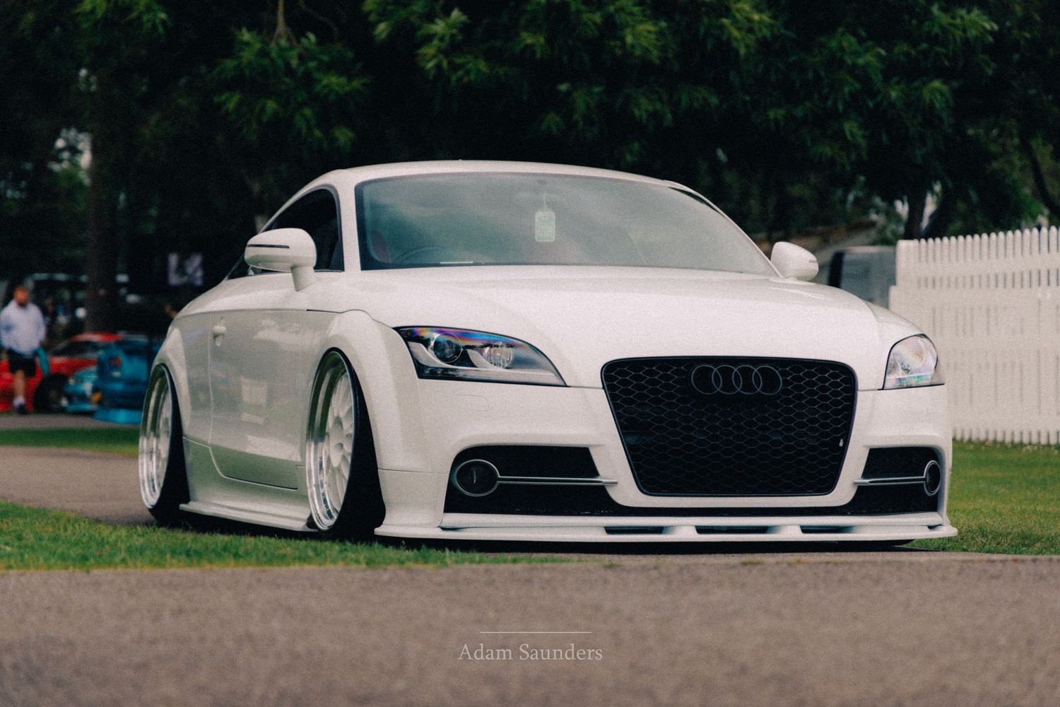 White Audi TT coupe with lowered suspension and custom wheels parked on grass at a car show.