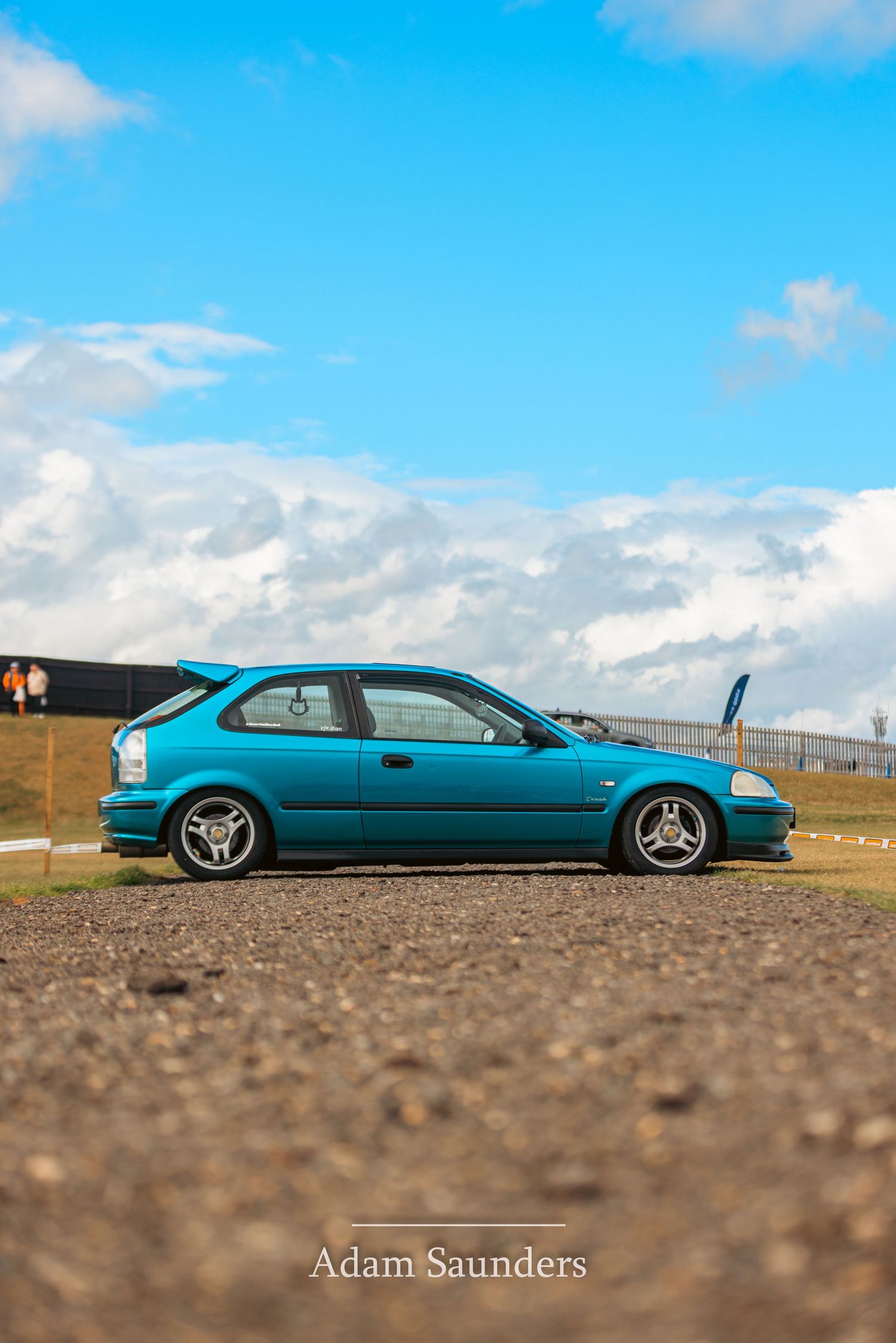 A teal Honda Civic EJ coupe sits low to the ground against a bright blue sky with white clouds.