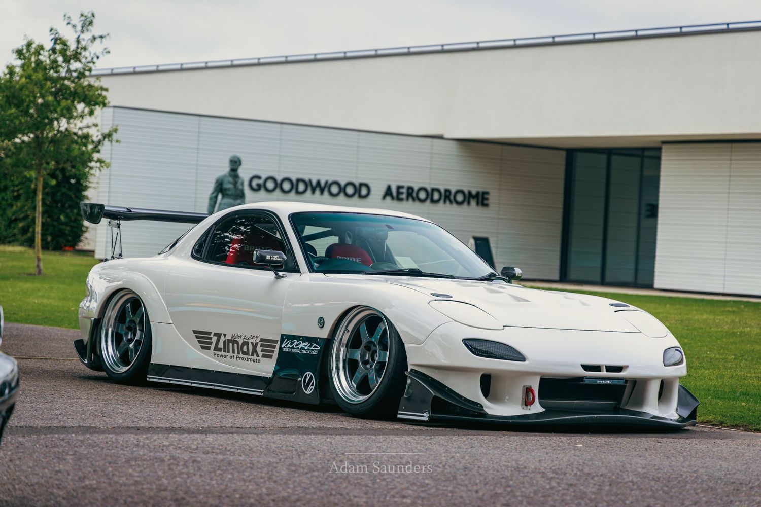 White Mazda RX-7 with wide body kit parked at Goodwood Aerodrome, featuring aggressive stance and custom wheels.
