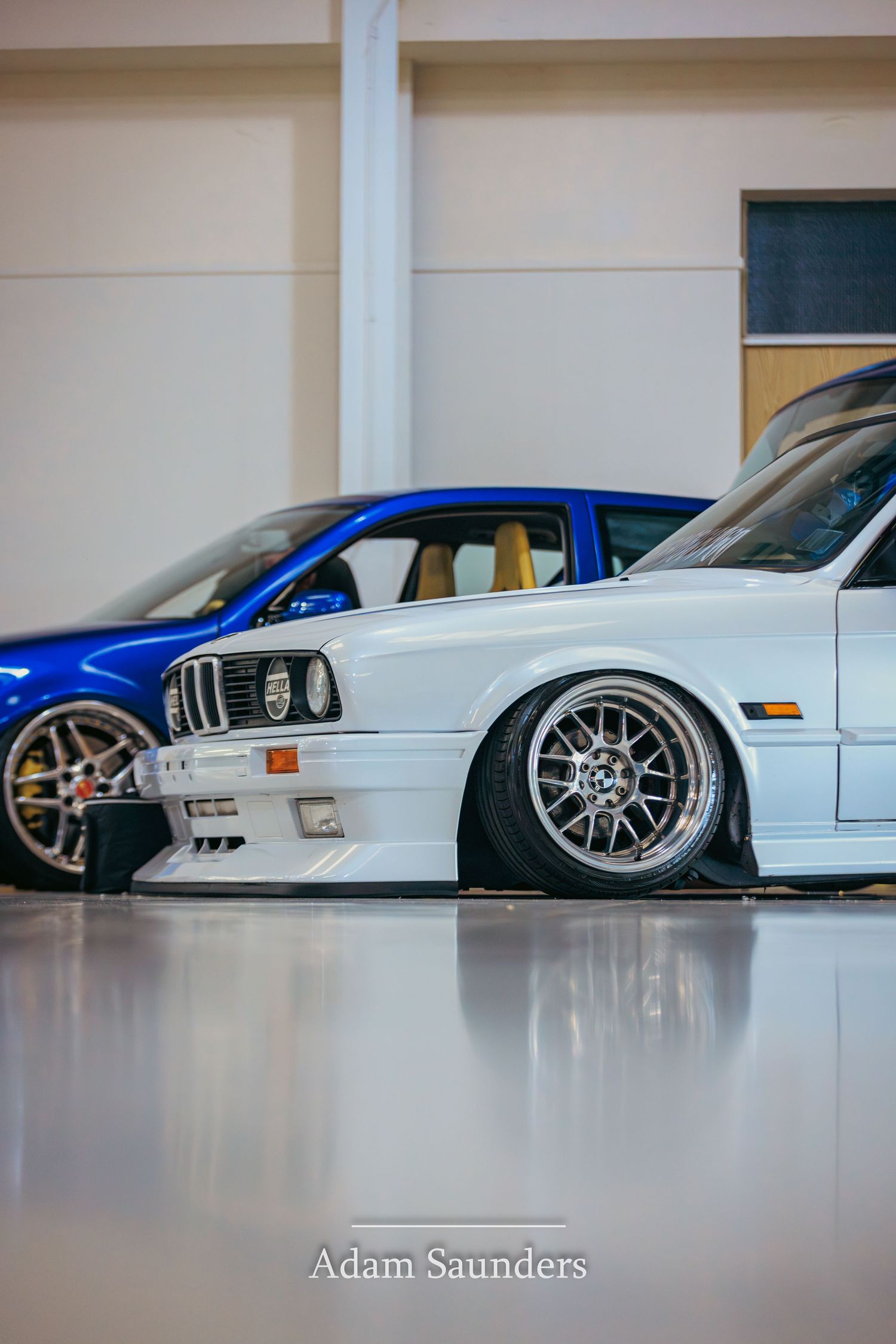 Modified white BMW E30 with BBS-style wheels and lowered suspension sits next to a blue car in an indoor space.