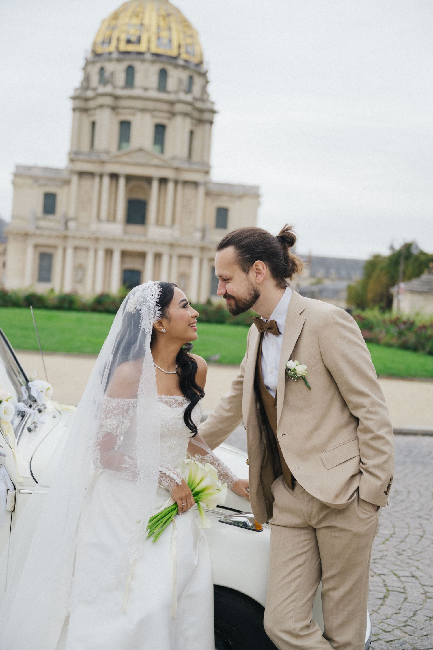 Les invalides wedding photoshoot with a white vintage car. The bride is wearing a white dress and the groom is wearing a beige suit.