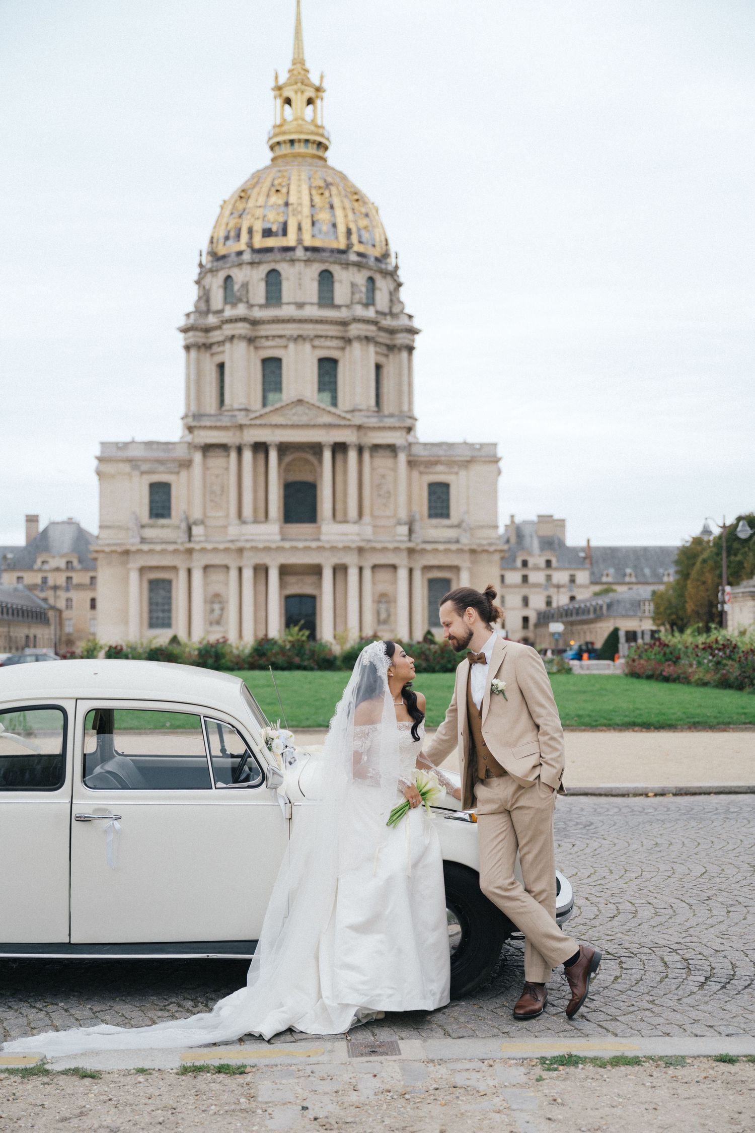 Les invalides wedding photoshoot with a white vintage car. The bride is wearing a white dress and the groom is wearing a beige suit.