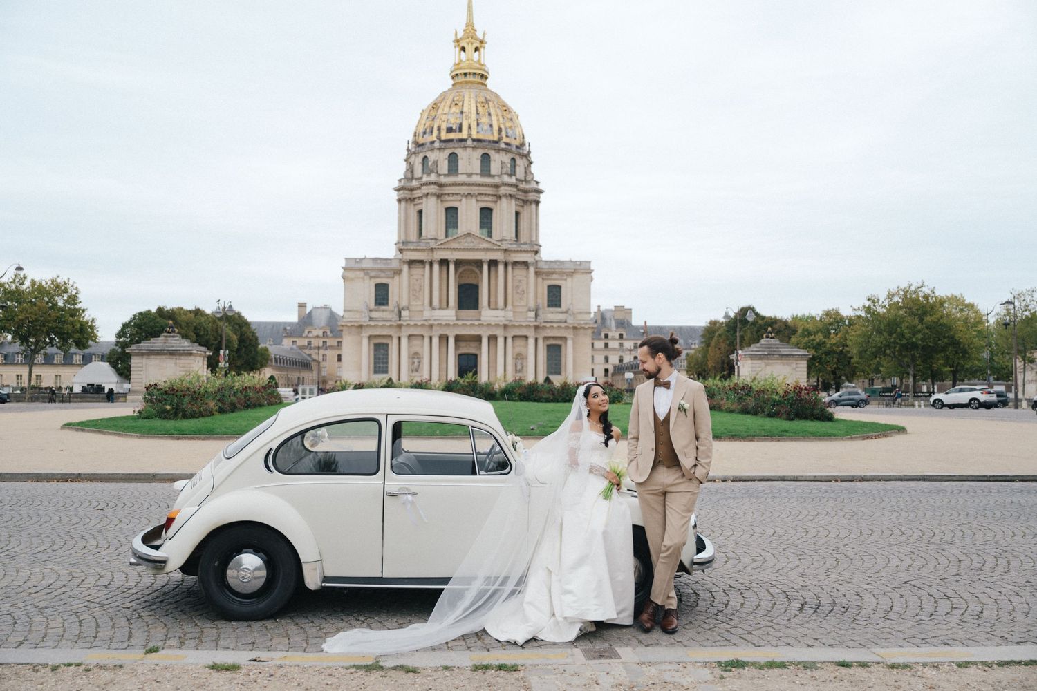 Les invalides wedding photoshoot with a white vintage car. The bride is wearing a white dress and the groom is wearing a beige suit.