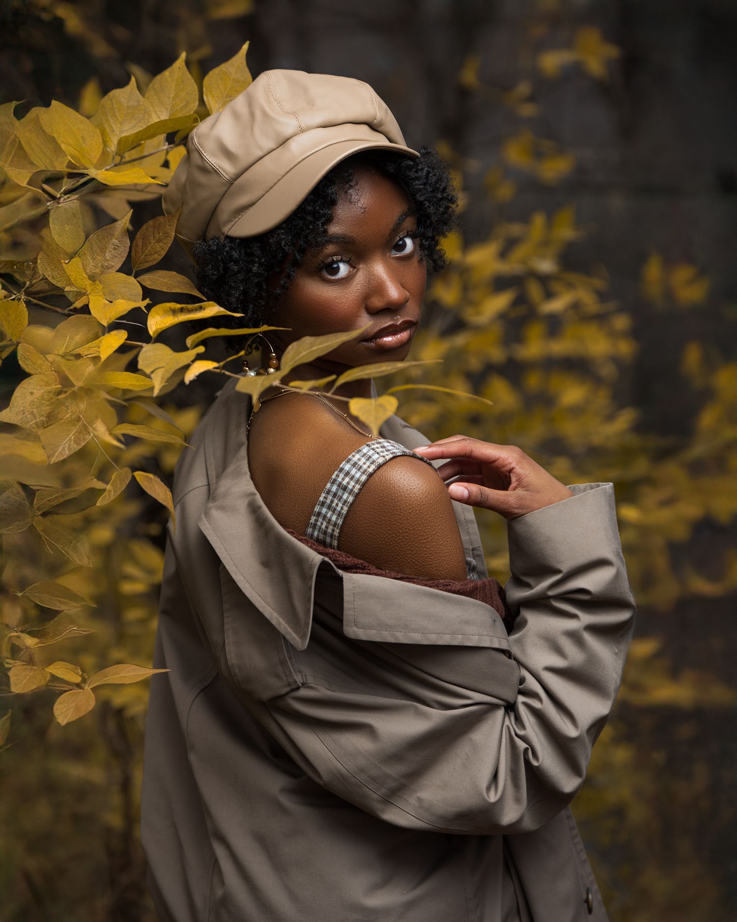 Artistic autumn portrait featuring a beige baseball cap and trench coat against yellow leaves in moody lighting.
