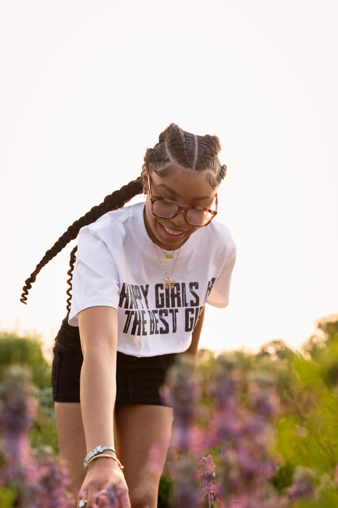 A person in glasses and a white t-shirt smiles while bending down among blooming purple flowers at sunset.