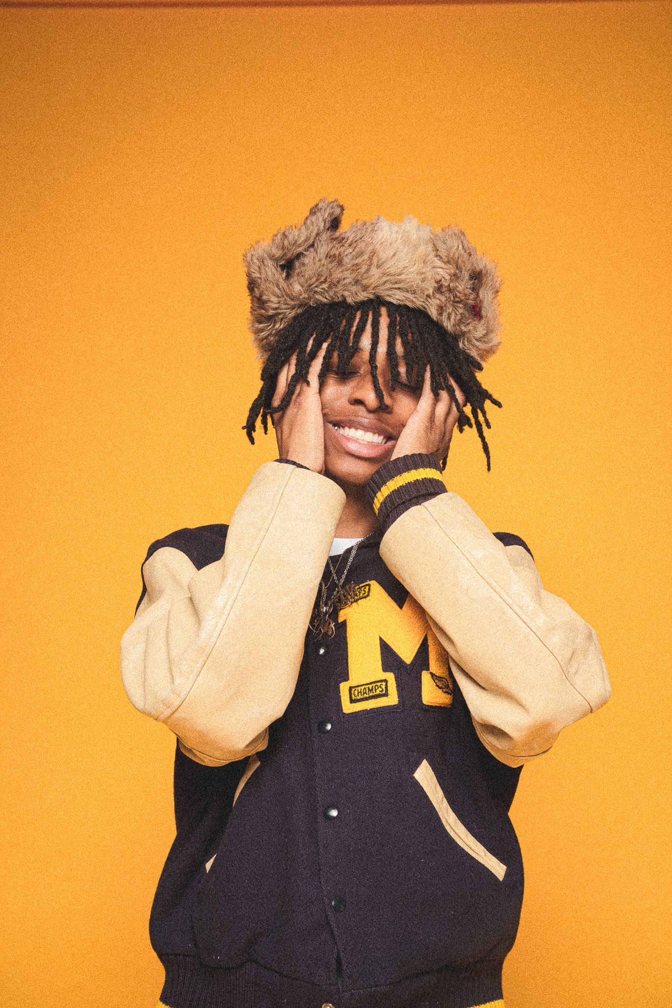 Fashion portrait with varsity jacket and furry hat against vibrant orange background.