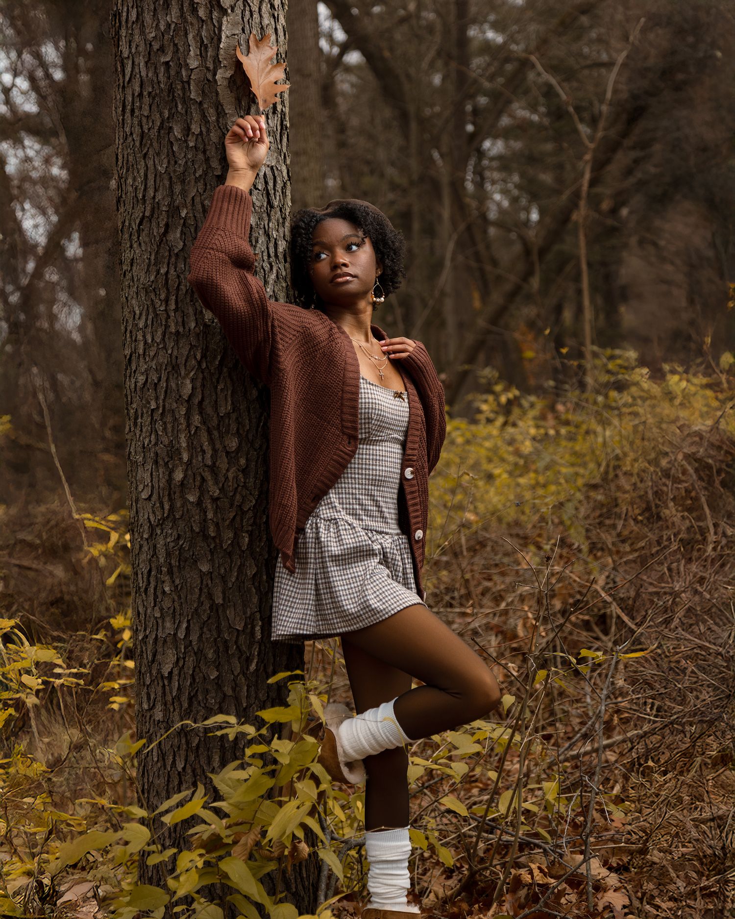 Person in brown cardigan and white dress poses against tree trunk in autumn forest with yellow foliage.