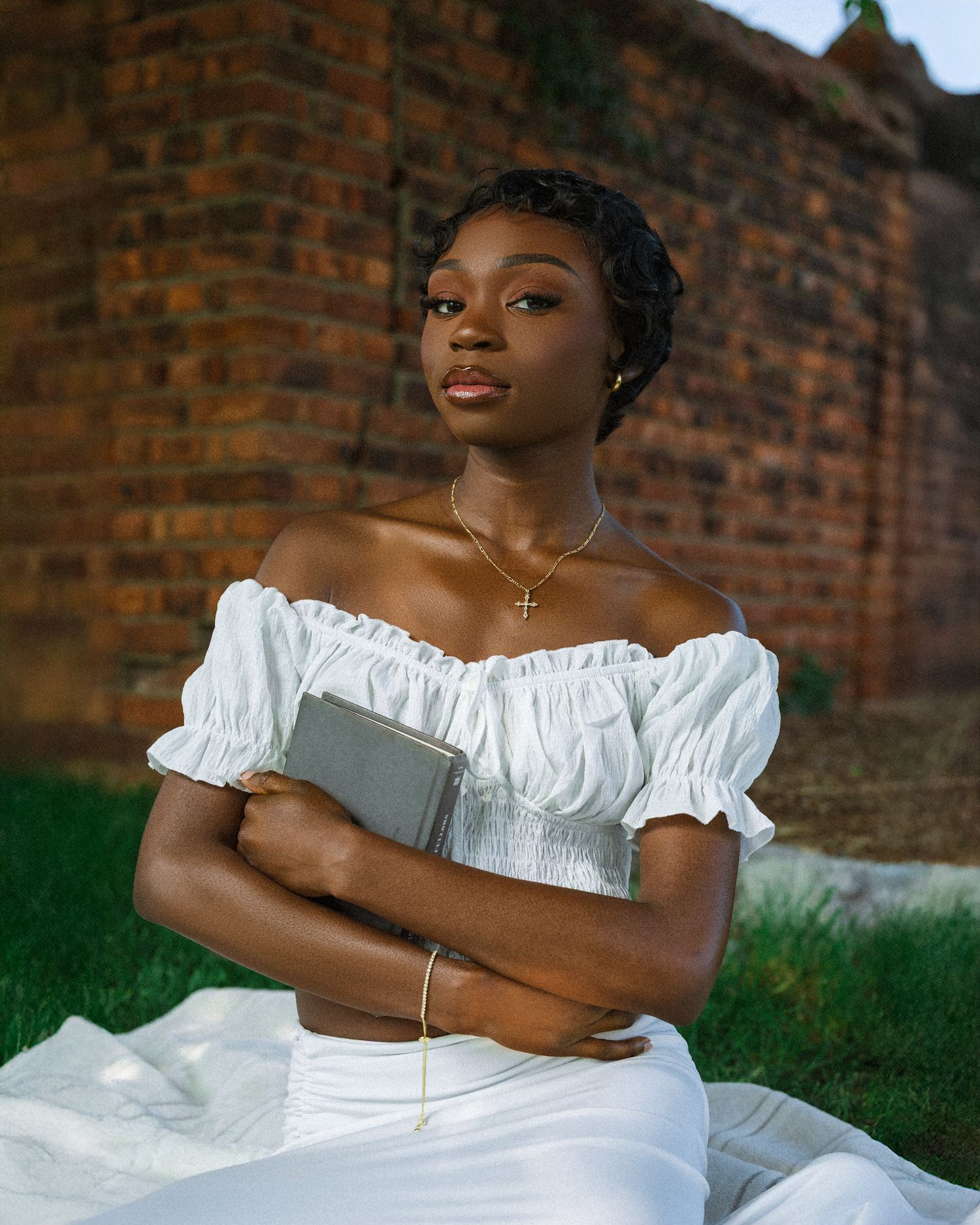 Person in white off-shoulder dress poses confidently against rustic brick wall backdrop during an outdoor photoshoot.