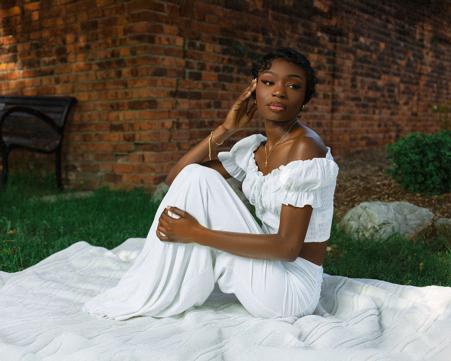 A woman in a white off-shoulder dress sits on a white blanket against a rustic brick wall backdrop.