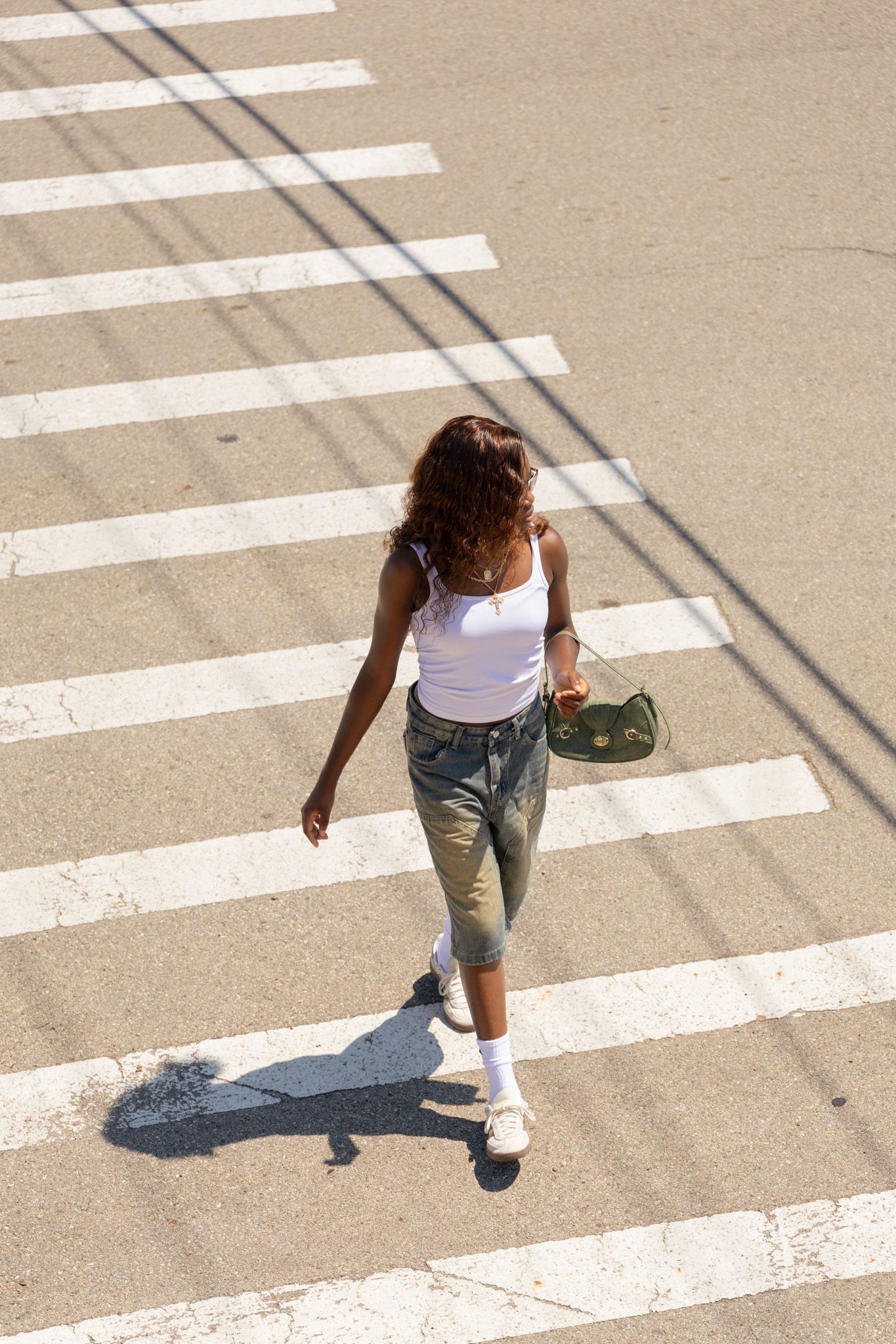 Person in white tank top and cropped jeans walking across a zebra crossing on a sunny day, casting long shadow.