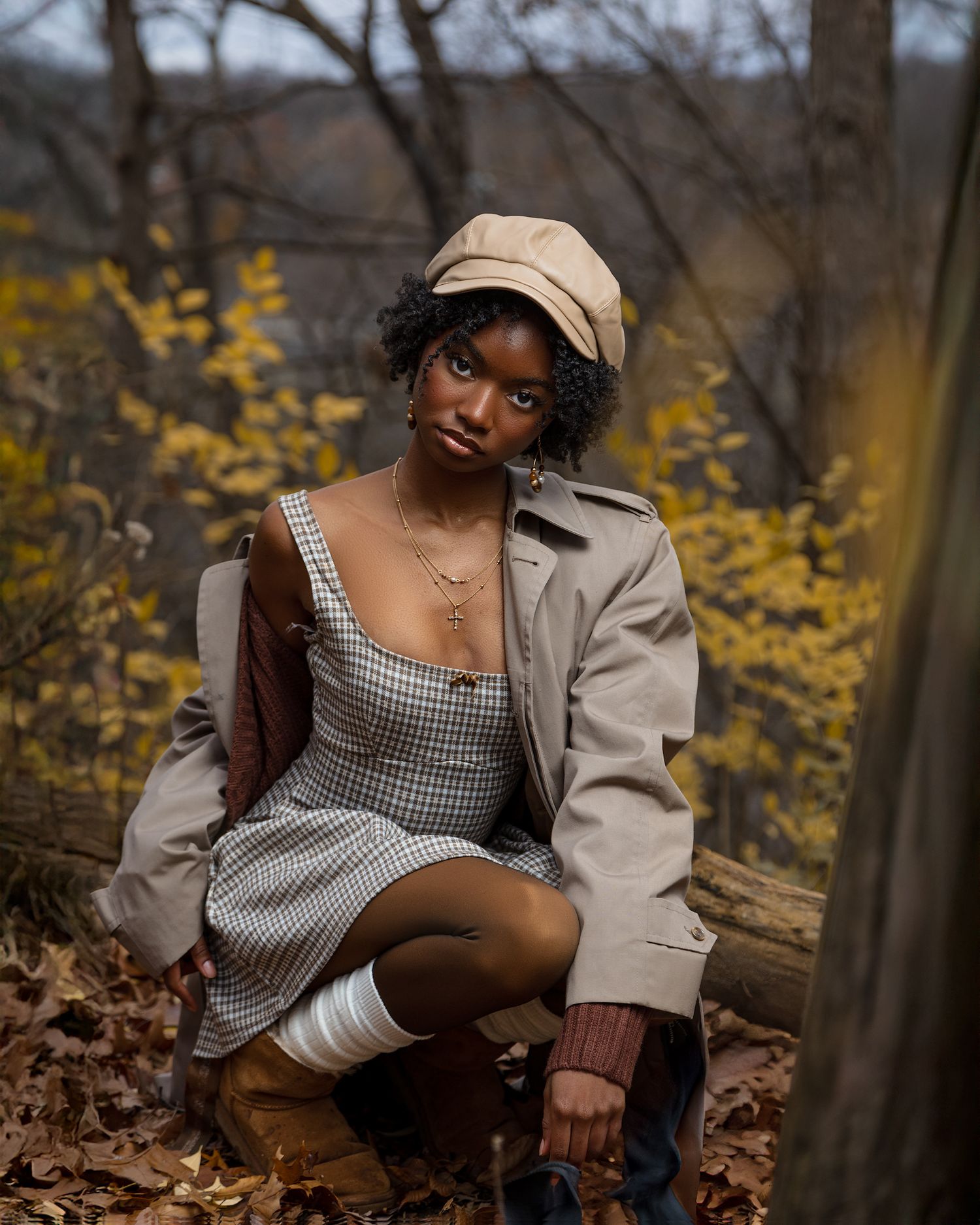 A person in a gingham top poses among fallen autumn leaves and yellow foliage in a moody forest setting.